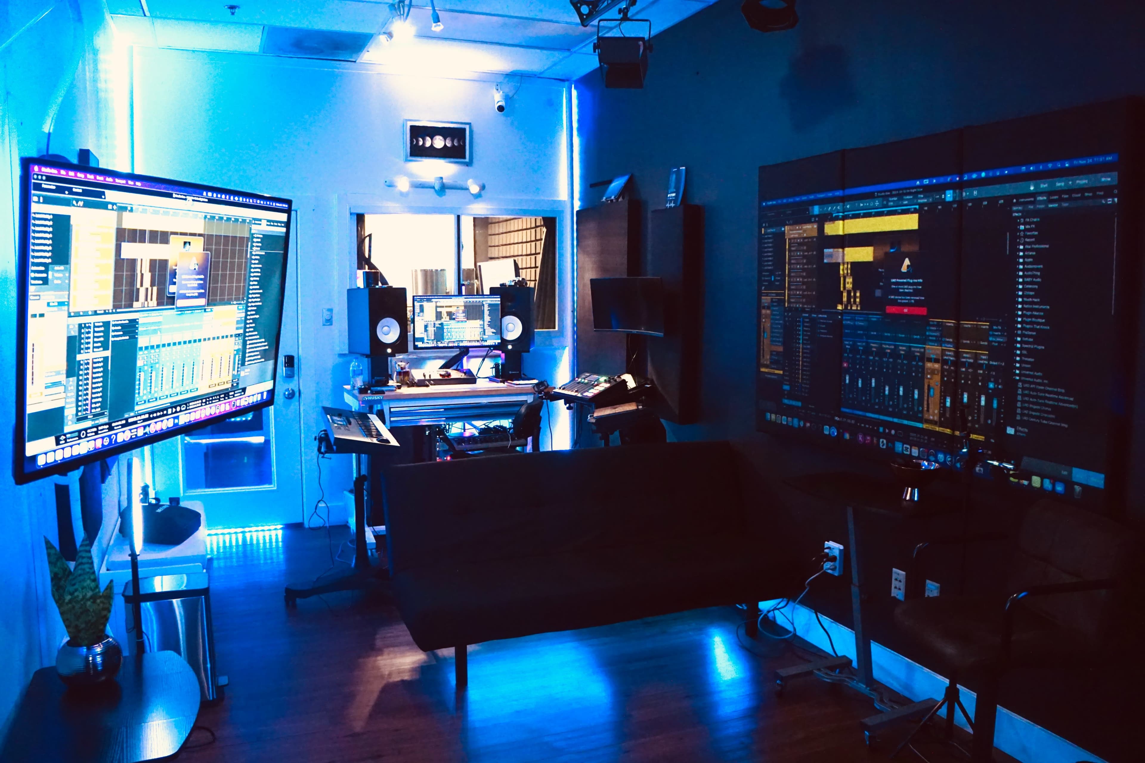 A recording studio with multiple large screens displaying audio software, a production desk, and a seating area.