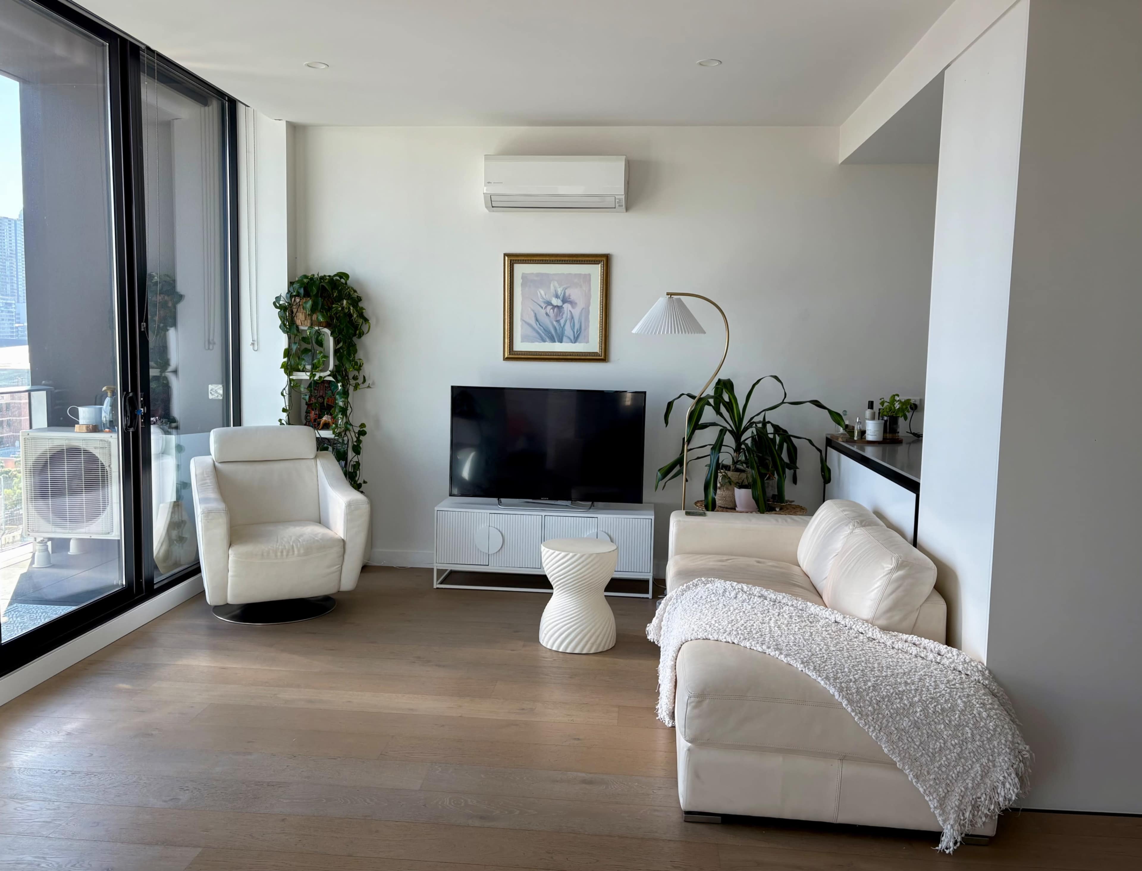 A modern living room features two white armchairs, a television on a sleek stand, a small side table, and large windows with a city view.