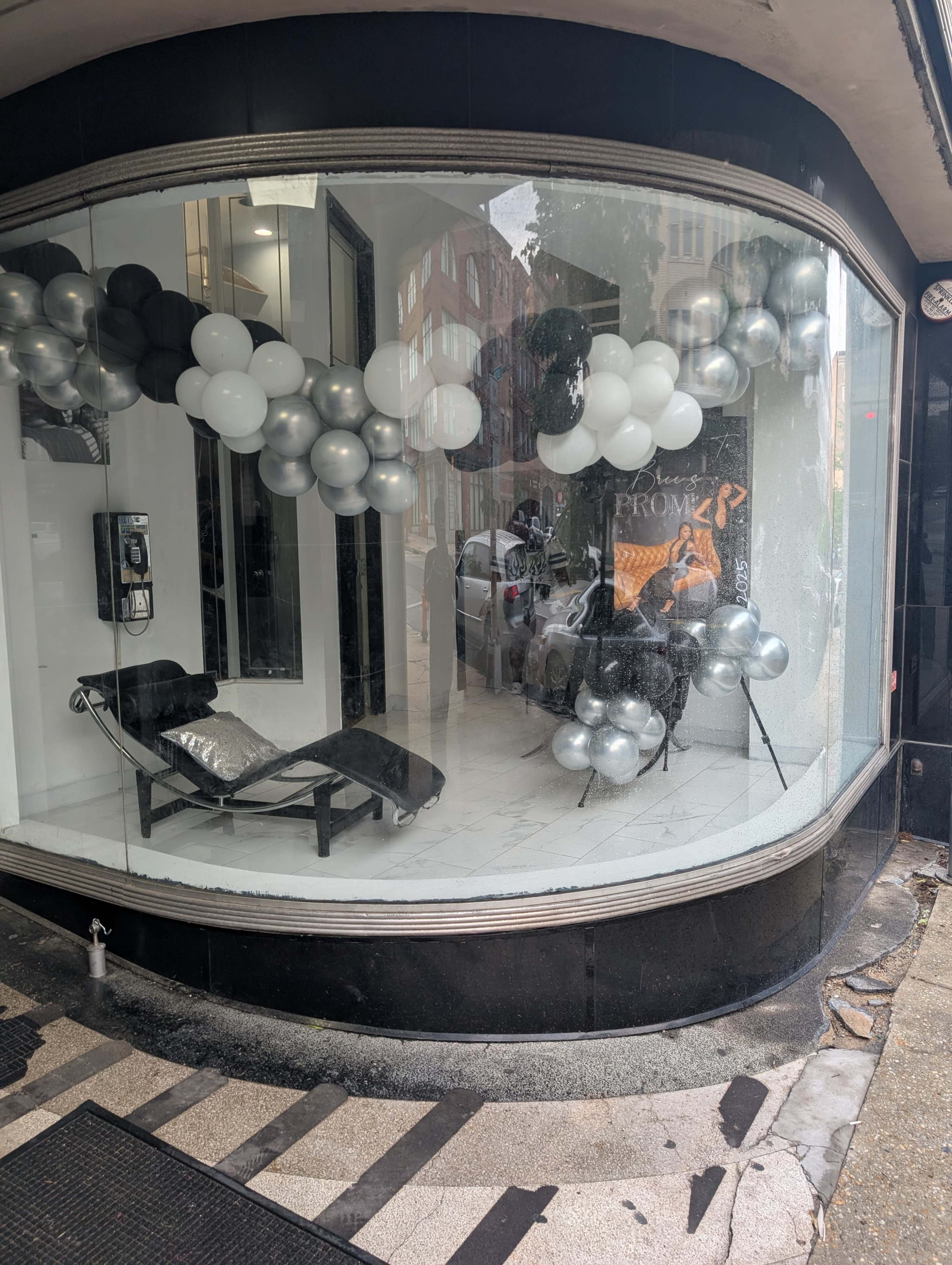 A storefront features a large display window decorated with black, silver, and white balloons, along with a lounge chair and promotional signage.