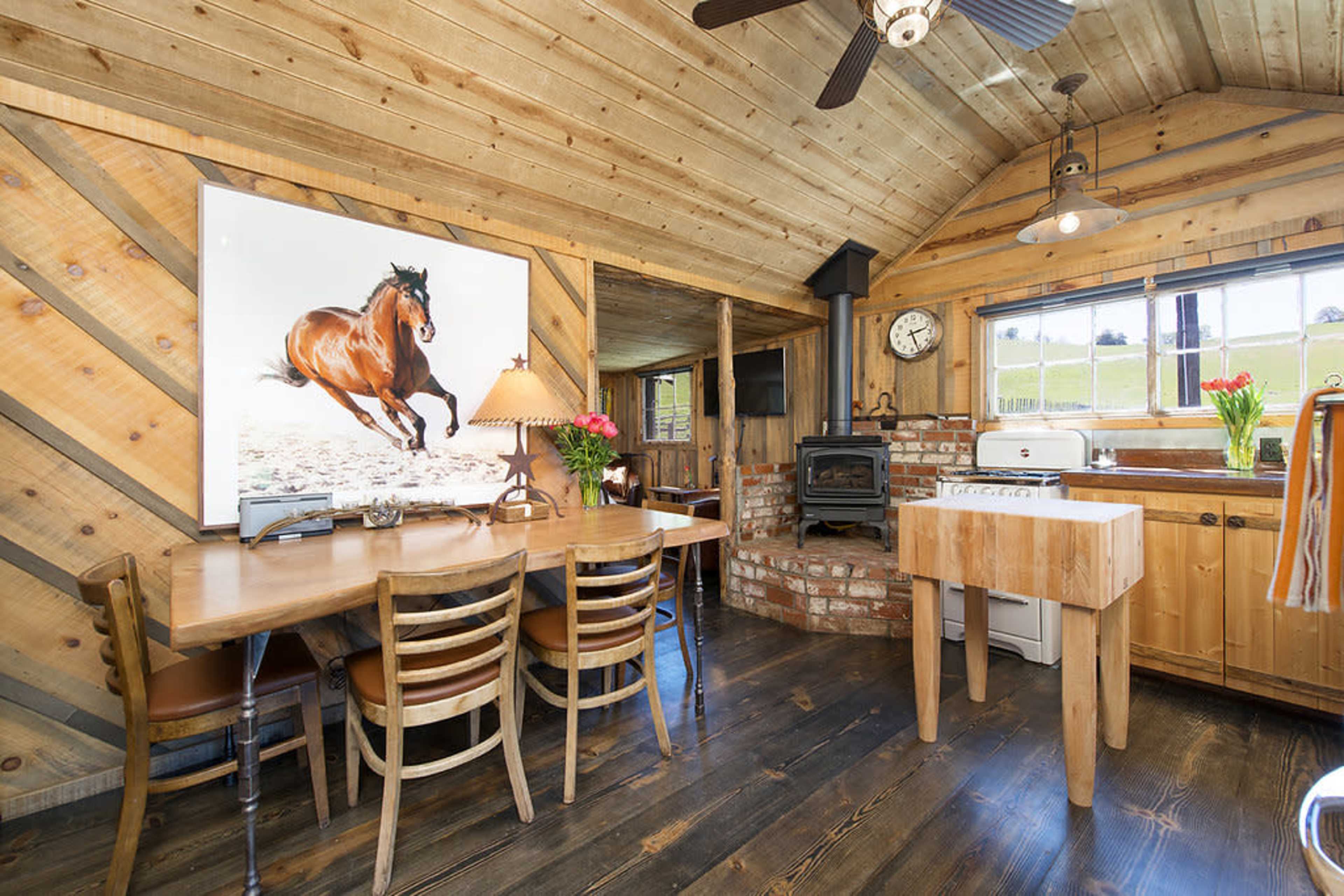 The image shows a rustic wooden interior of a cabin featuring a dining area with a table and chairs, a kitchen area with a stove, and a large framed picture of a horse on the wall.