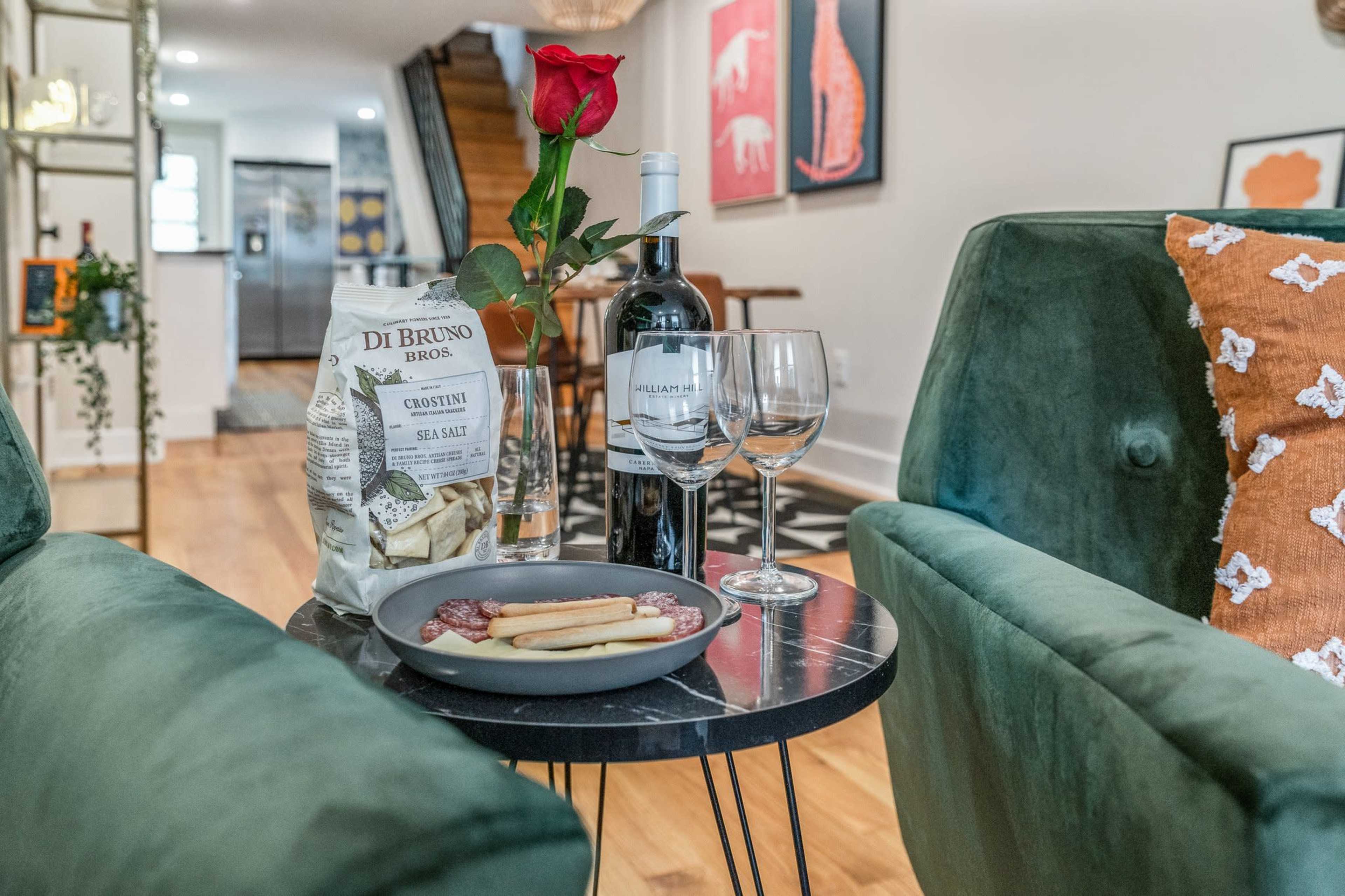 A cozy living area features two green chairs, a small table with a rose, a bottle of wine, wine glasses, crackers, and a plate with sliced cheese and jam.