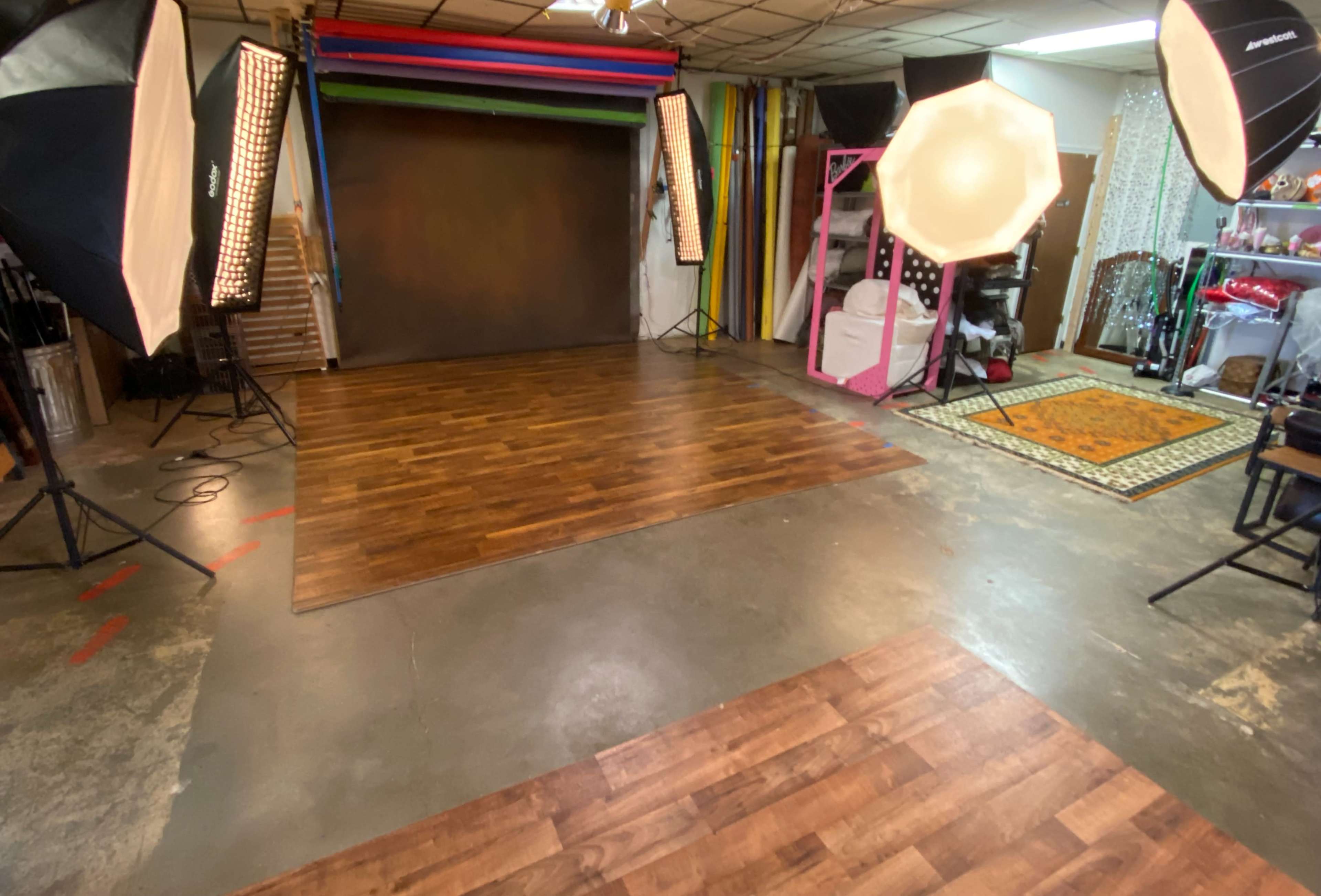 The image shows a photography studio with a wooden floor, backdrop setup, and various lighting equipment arranged in the space.