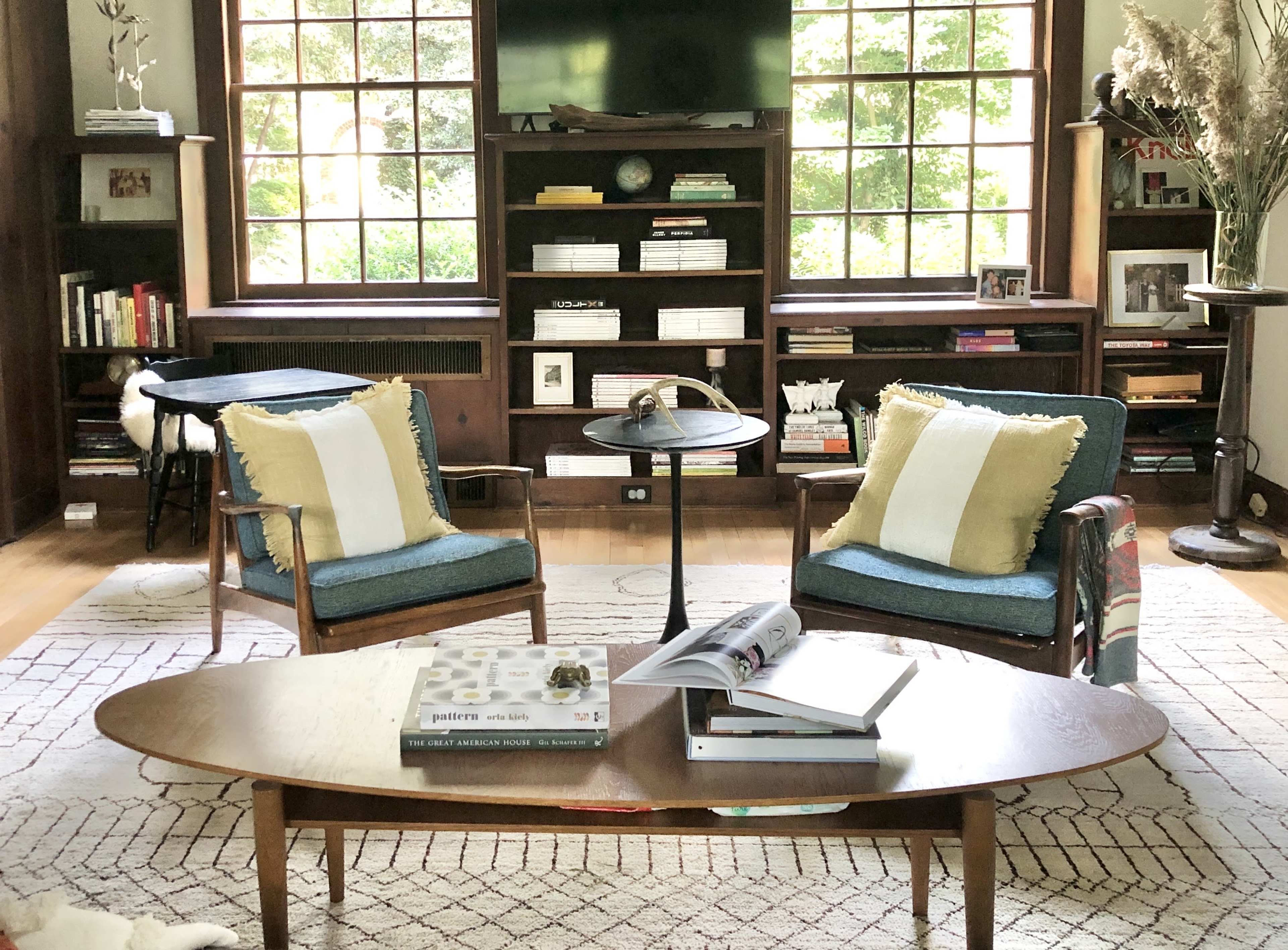 A living room features two chairs with cushions, a coffee table with books, and a large window overlooking greenery.