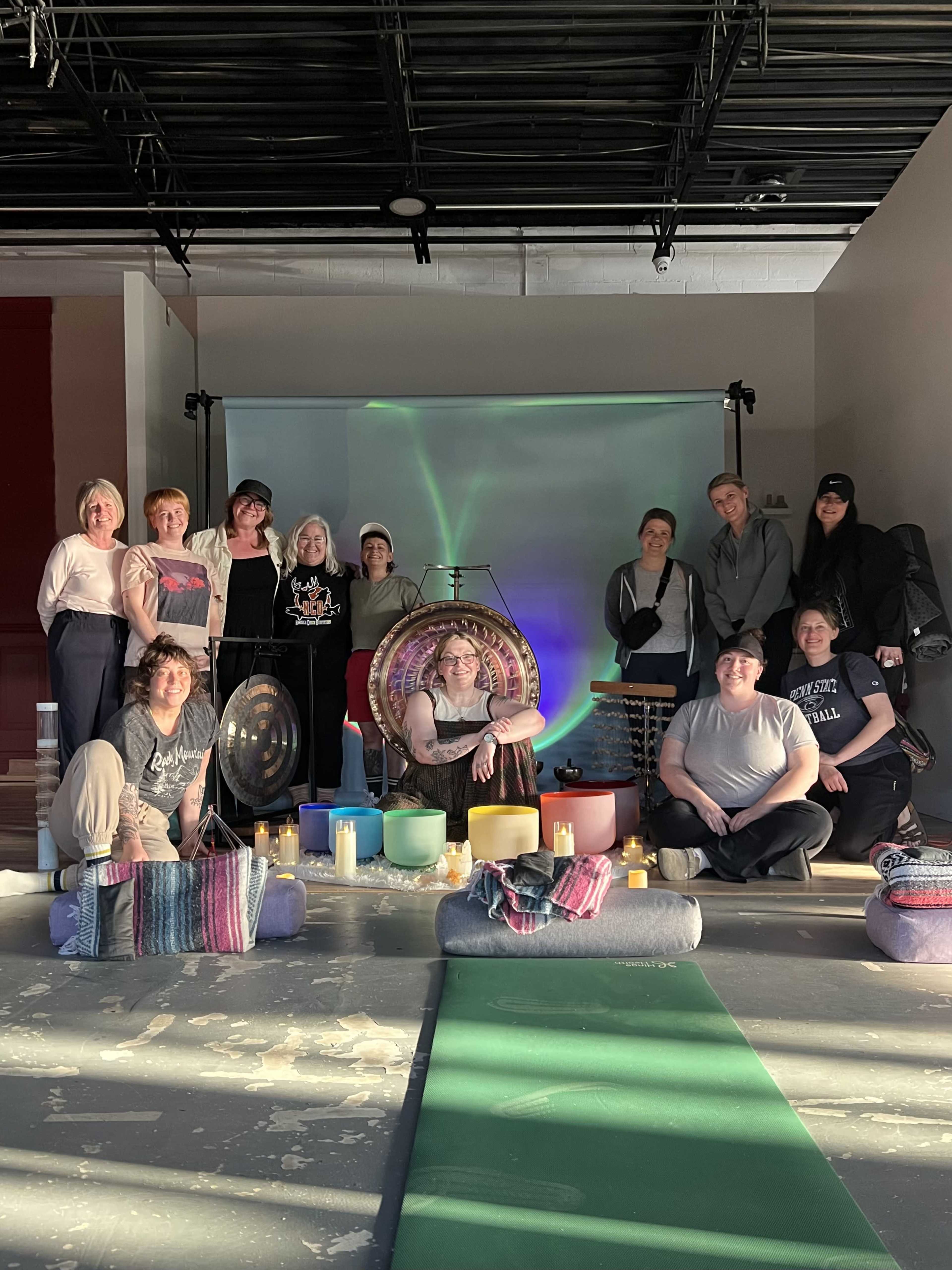 A group of women poses together in a yoga studio, surrounded by meditation props and ambient lighting.