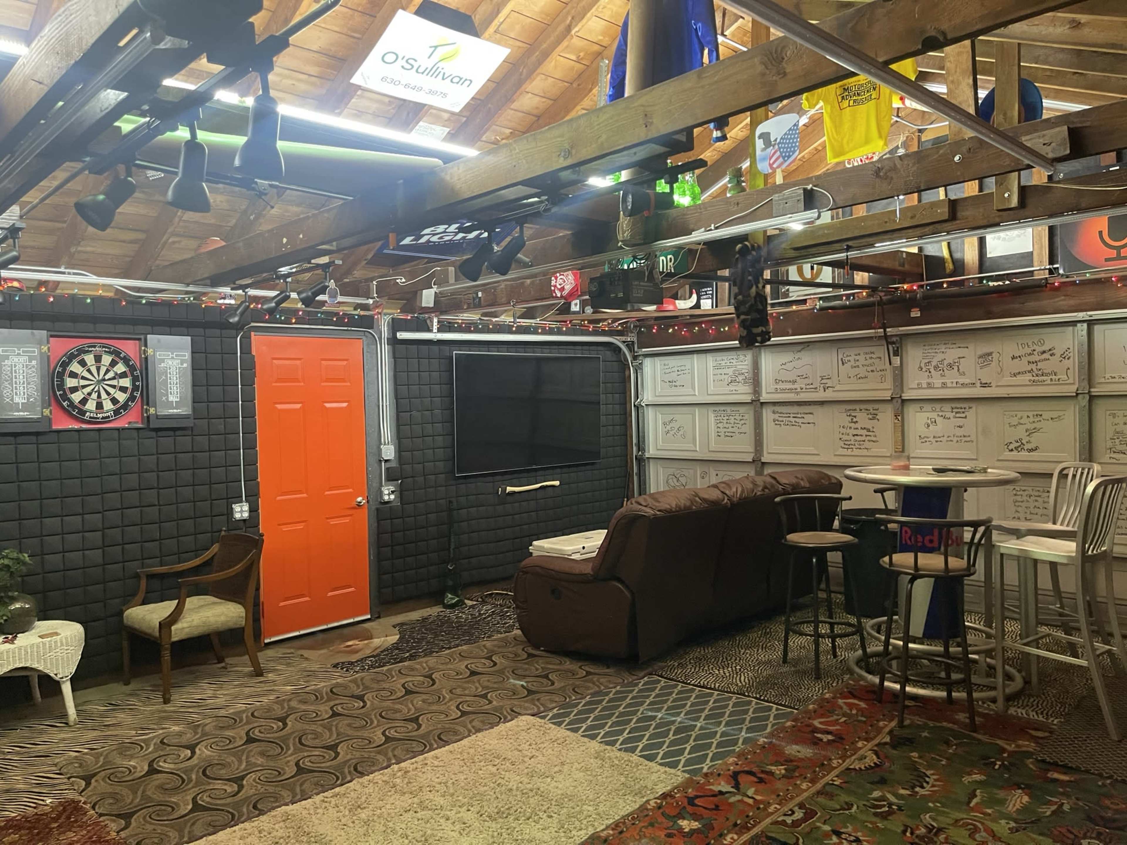 A cozy game room with an orange door, a dartboard, a television on the wall, and various seating arrangements under a wooden ceiling adorned with sports memorabilia.