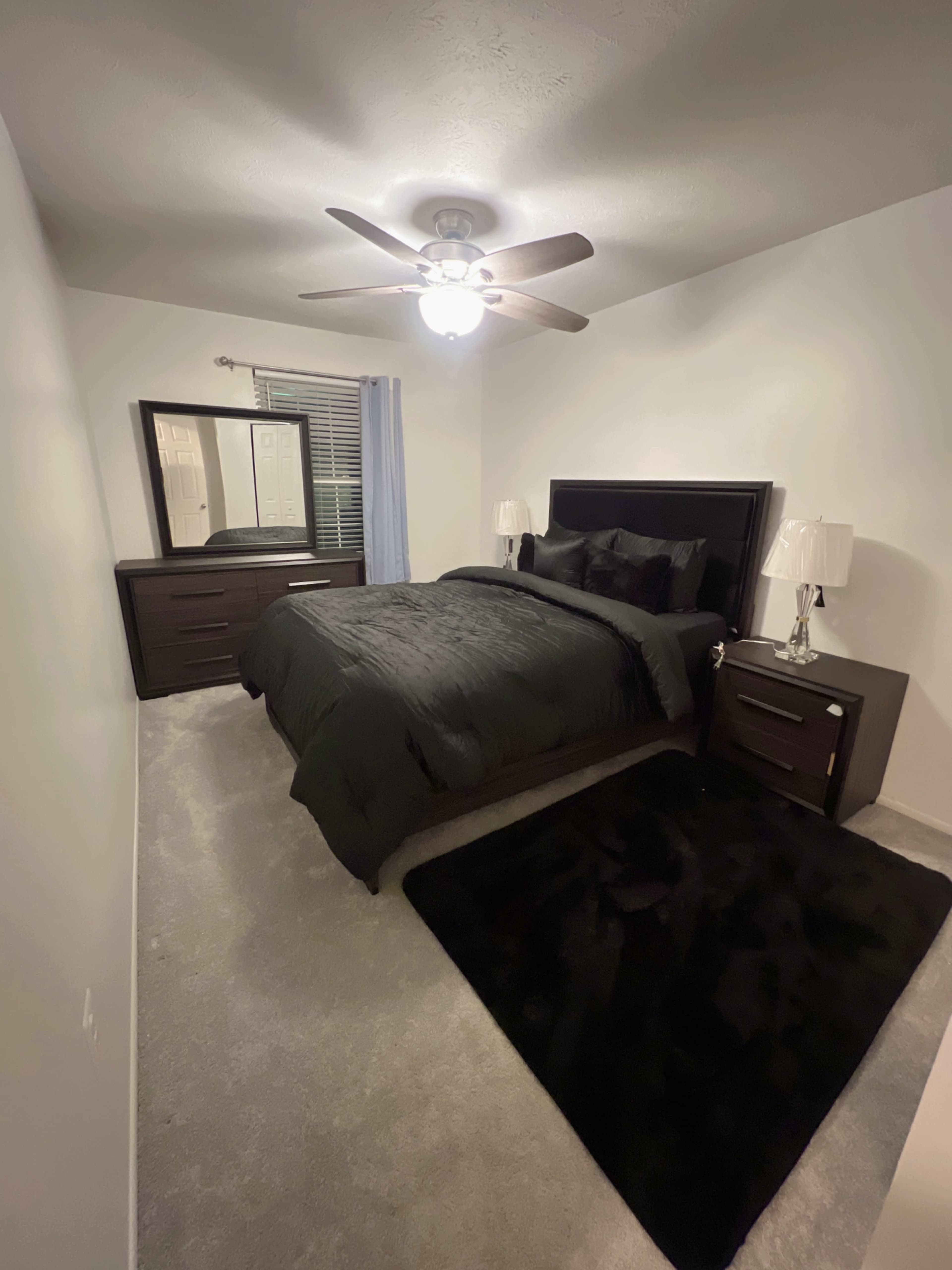 The image shows a neatly arranged bedroom featuring a bed with black bedding, matching nightstands, a dresser, and a ceiling fan.