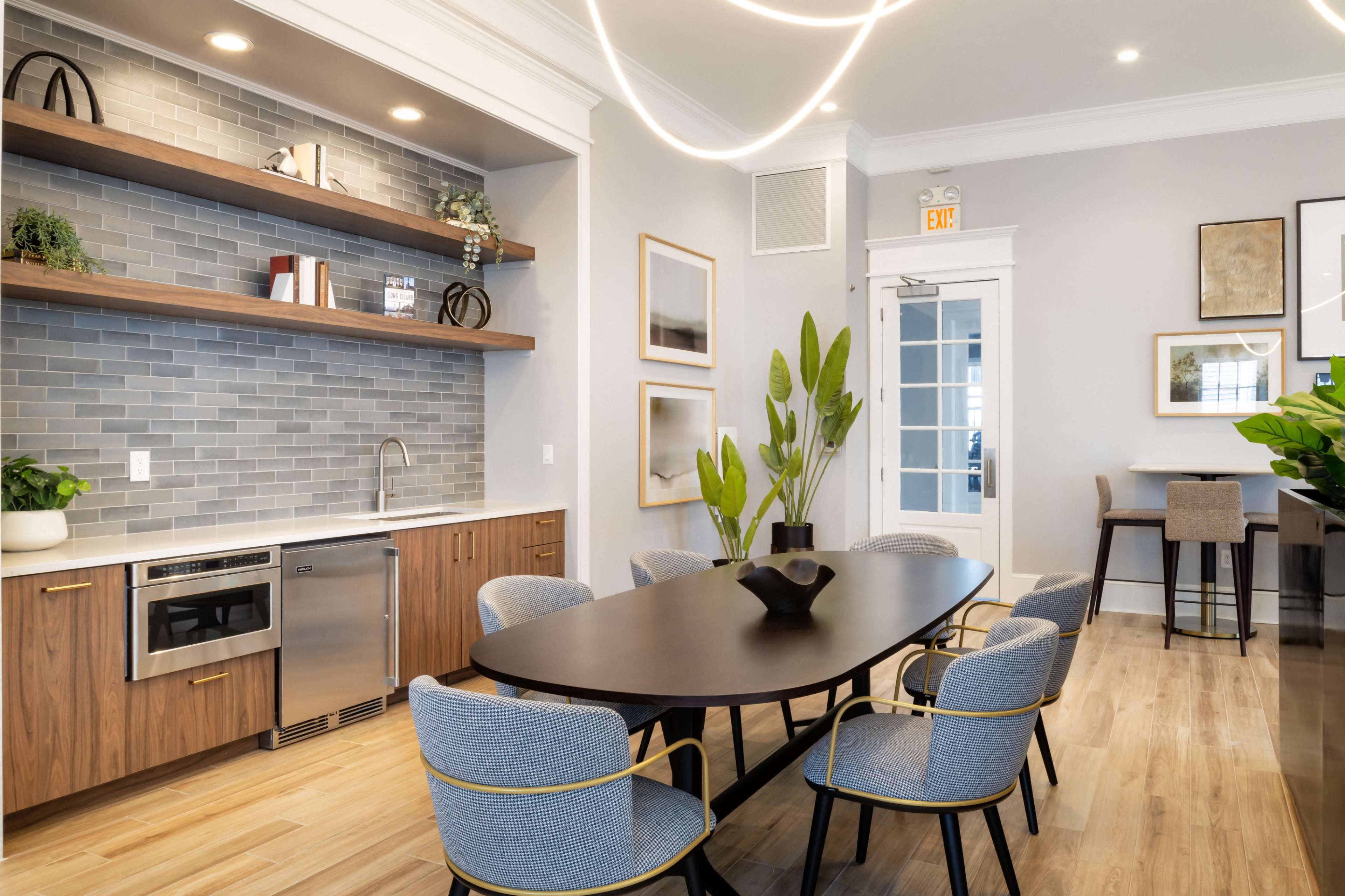 The image shows a modern kitchen and dining area featuring a dark wooden table surrounded by light gray chairs, a kitchenette with stainless steel appliances, and decorative plants.