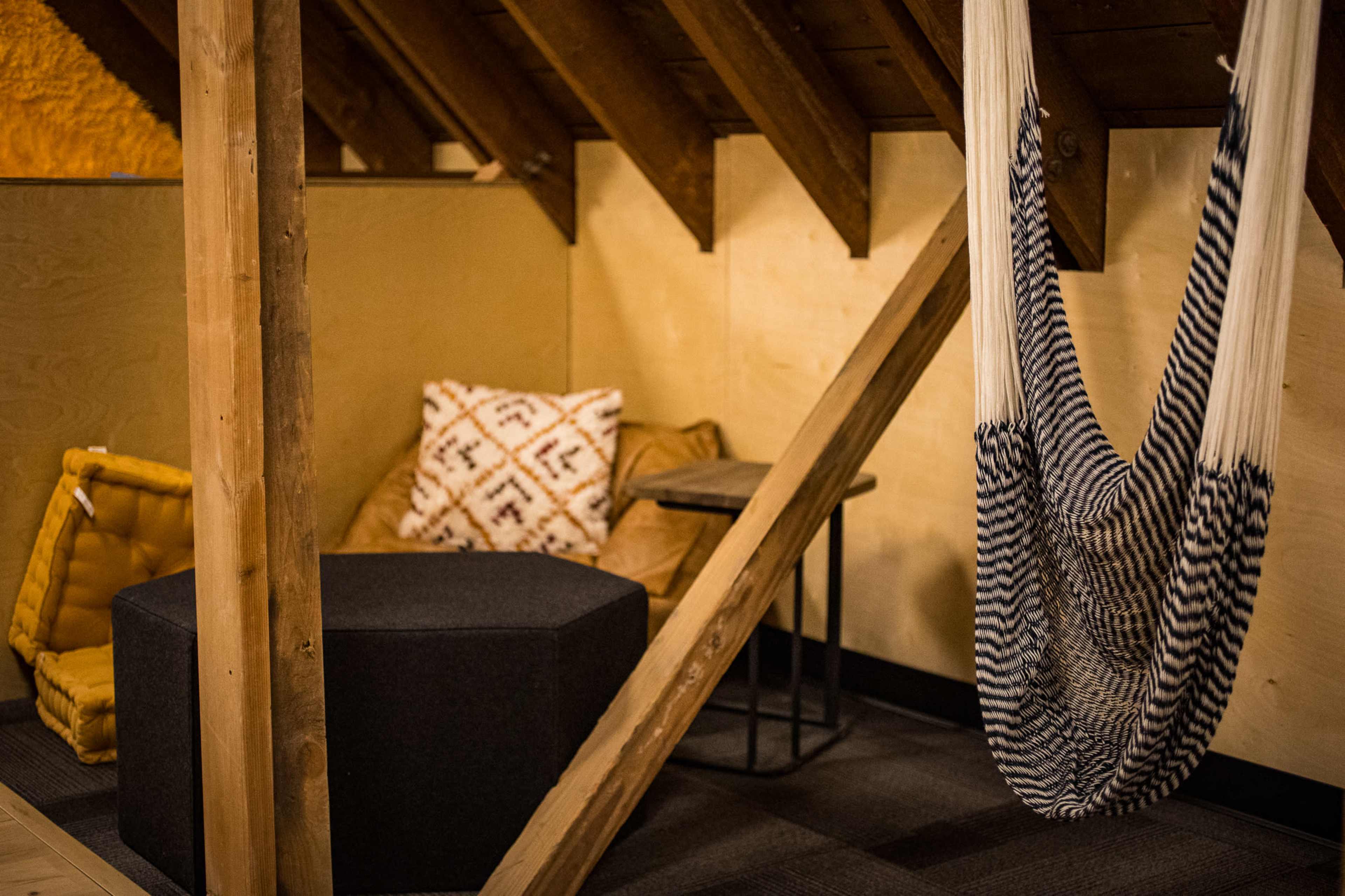 The image shows a cozy attic space featuring a hammock, a bean bag chair, a black ottoman, and wooden beams.