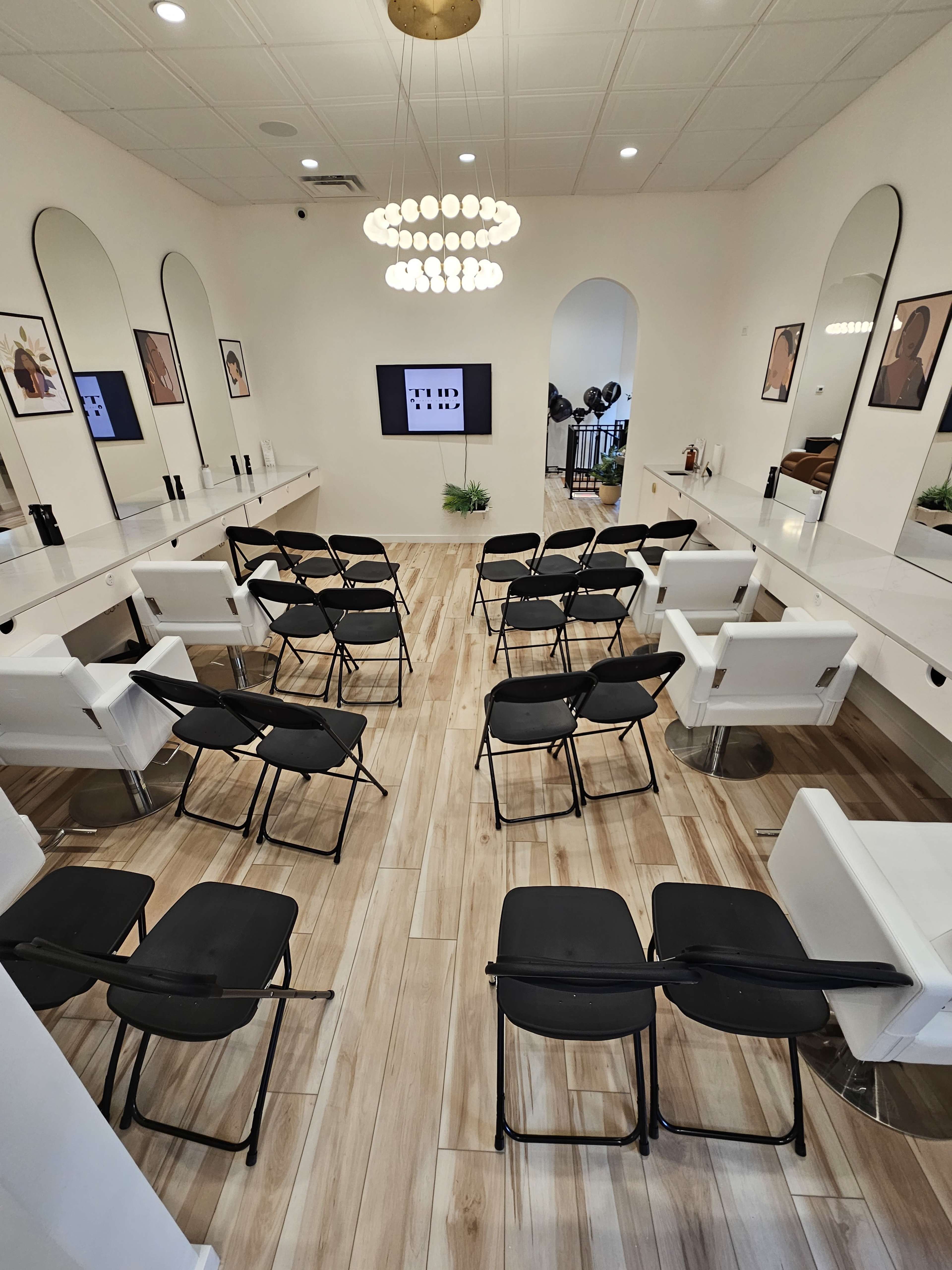 A modern salon interior with several black and white chairs arranged in rows facing a display screen, surrounded by mirrors and framed artwork on the walls.