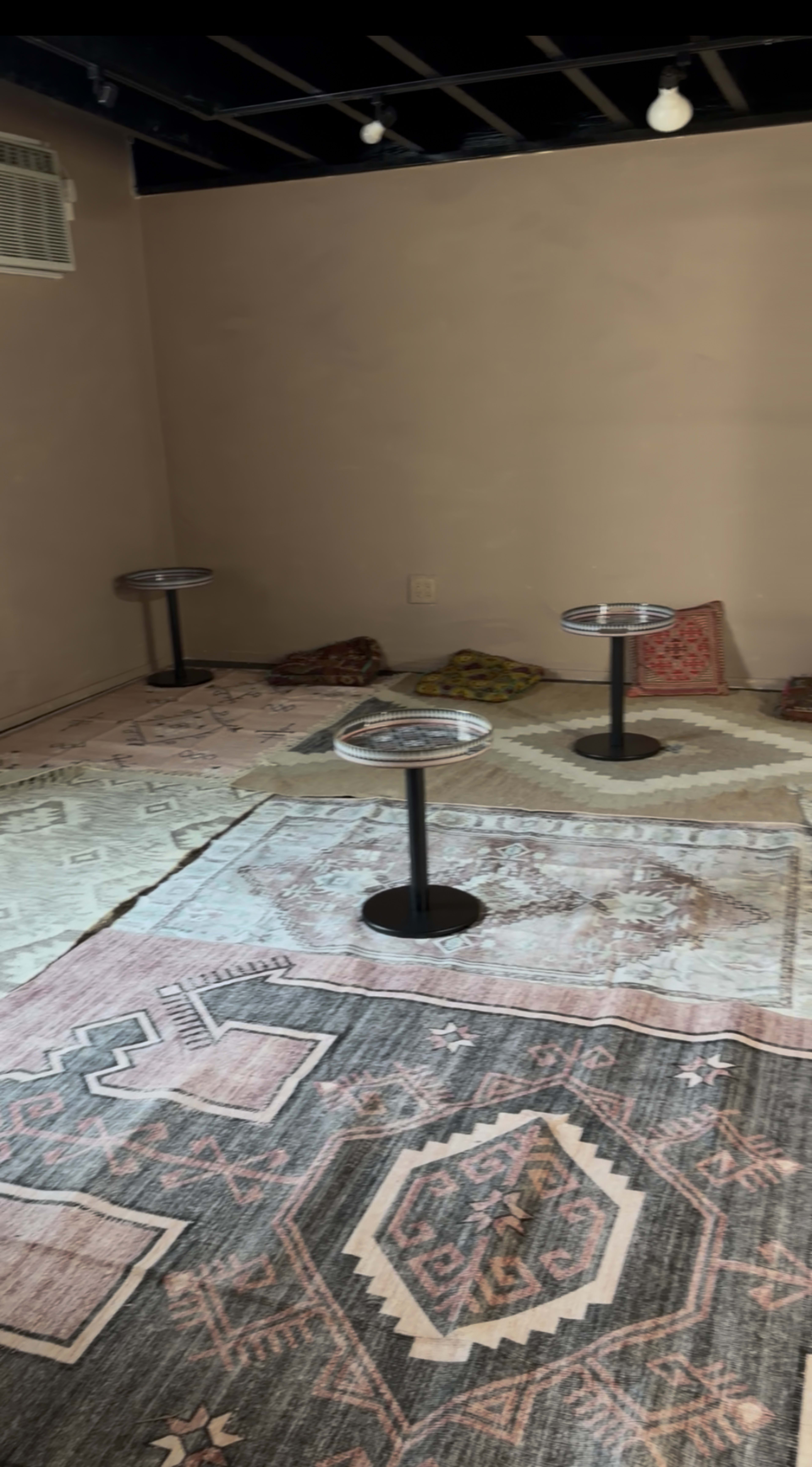 The image shows a sparsely furnished room with three small tables on the floor and decorative rugs spread out.