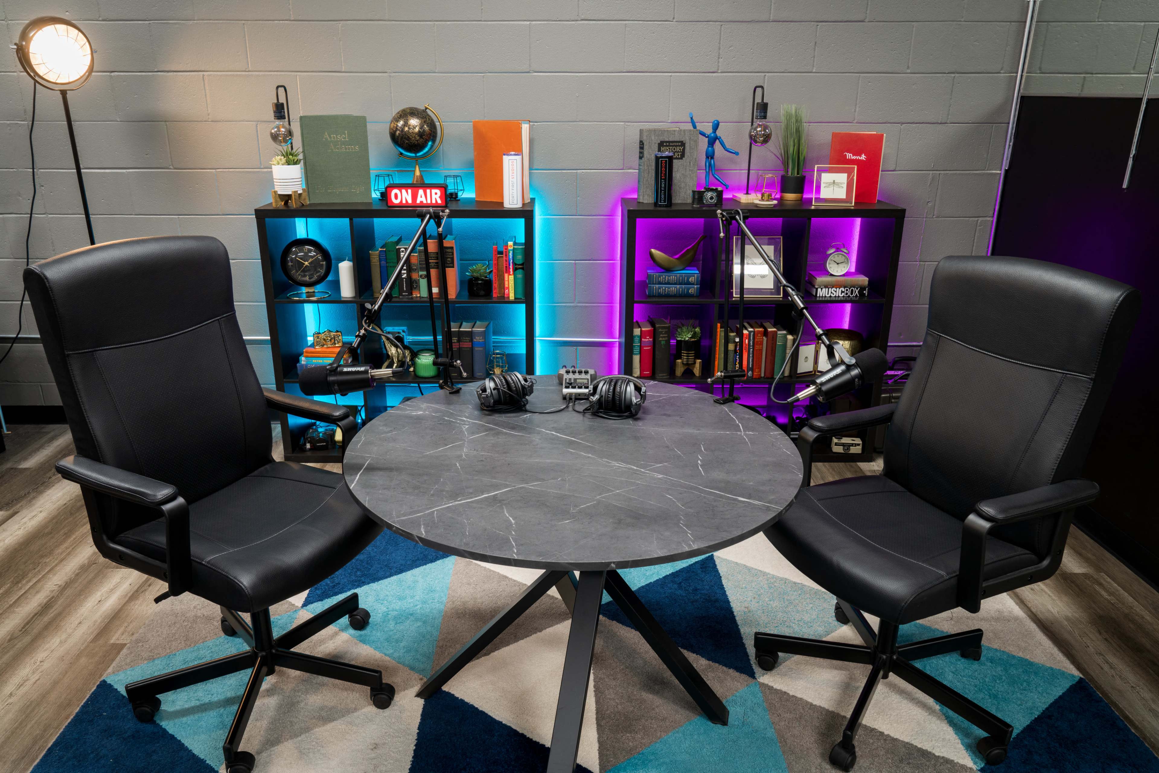 A round table is positioned between two black office chairs, surrounded by shelves with books and decorative items, illuminated by colored lights.