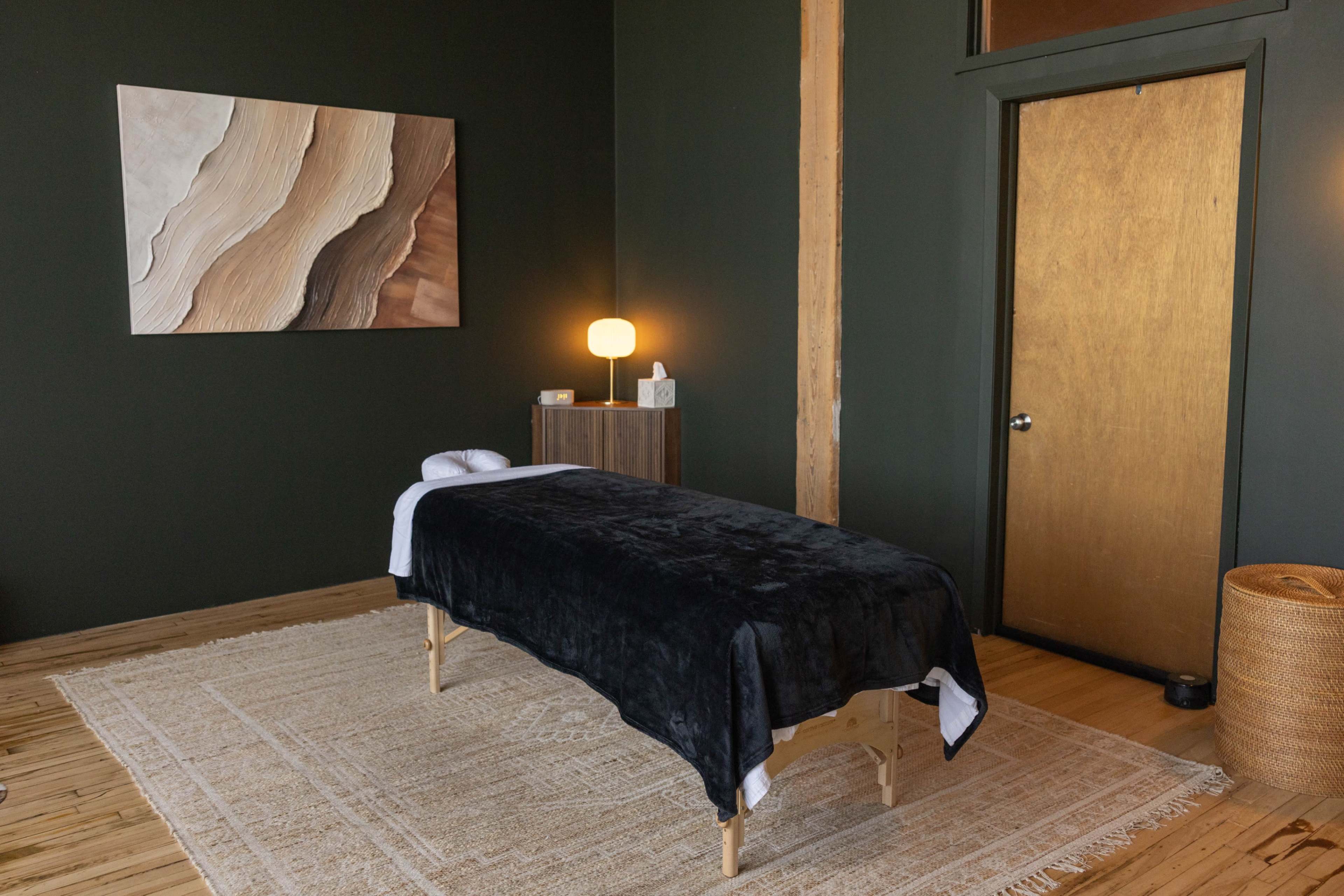 The image shows a spa treatment room featuring a massage table covered with a dark blanket, a wooden cabinet, a lamp, and a large artwork on the wall.