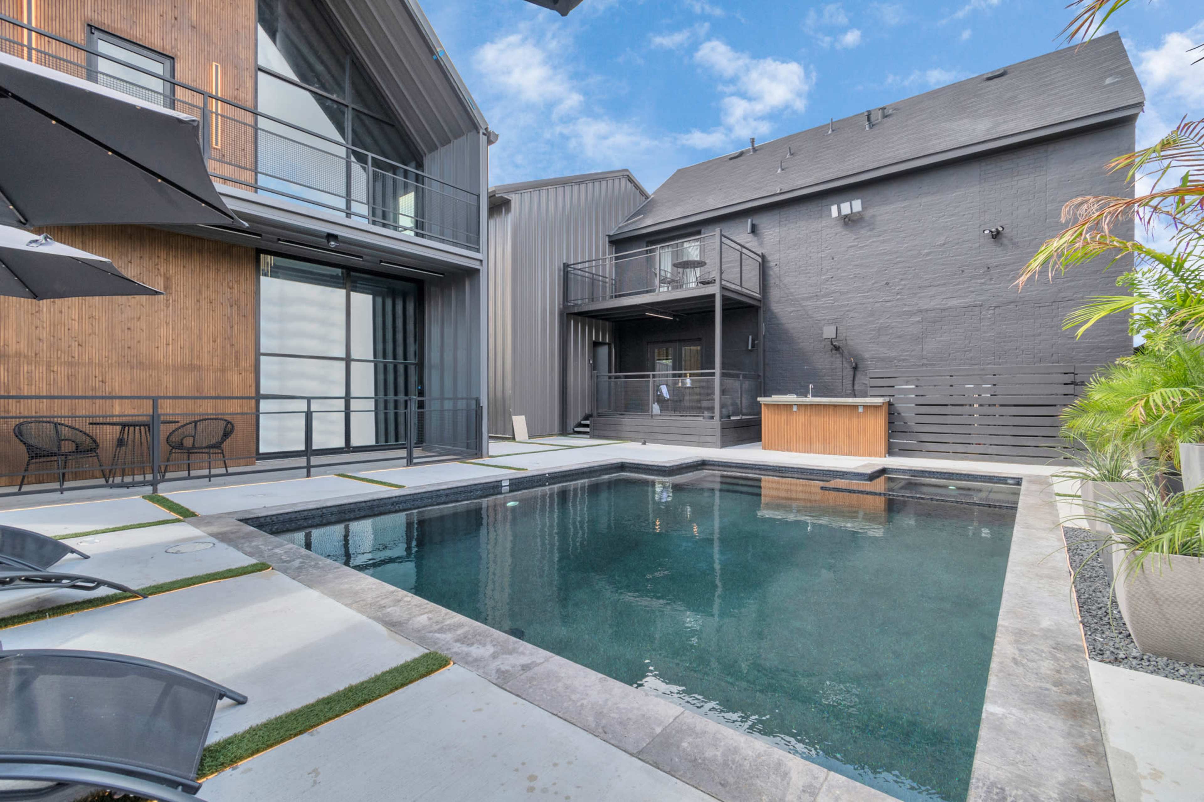 A modern outdoor space featuring a rectangular pool surrounded by concrete and grass, with adjacent seating areas and two structures in the background.