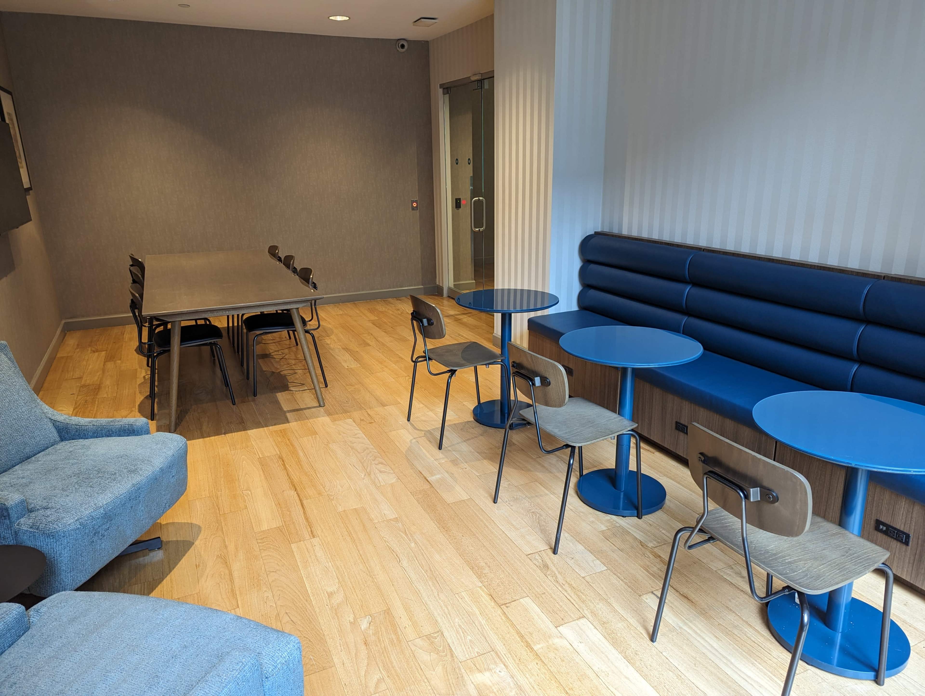 A spacious room with a mix of seating options, including blue tables and chairs, a long table, and a cozy seating area in a light-colored wood floor setting.