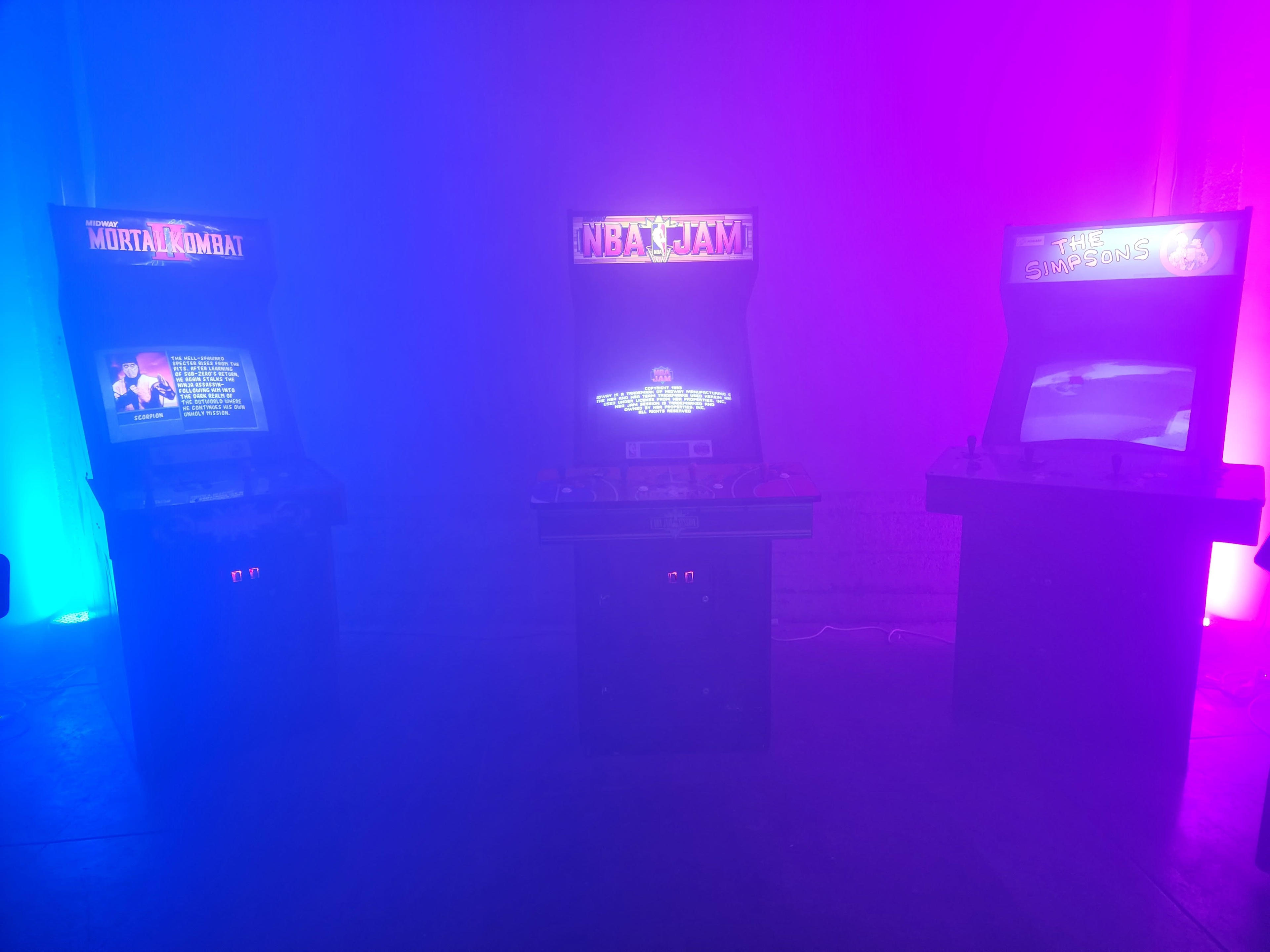 Three arcade game machines illuminated by purple and blue lighting in a foggy environment.