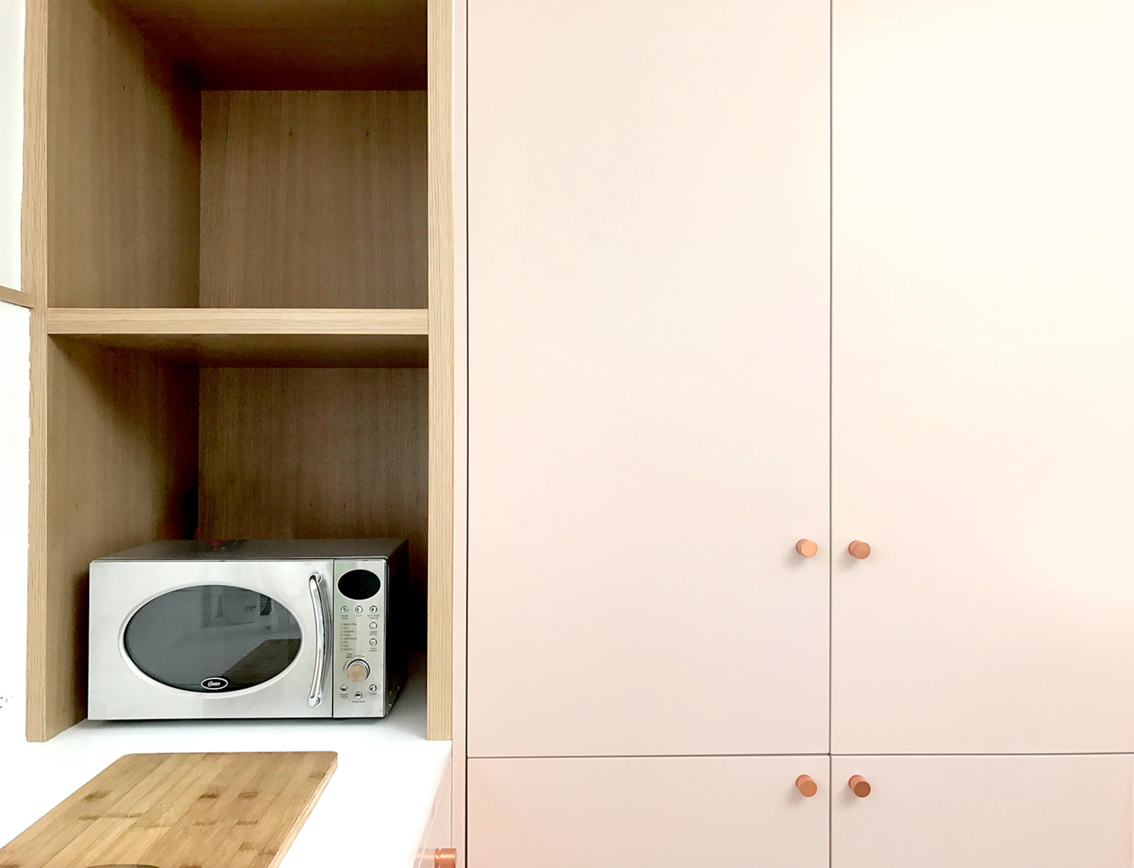 A white kitchen cabinet with a microwave placed on a shelf inside it, alongside a wooden cutting board on the countertop.