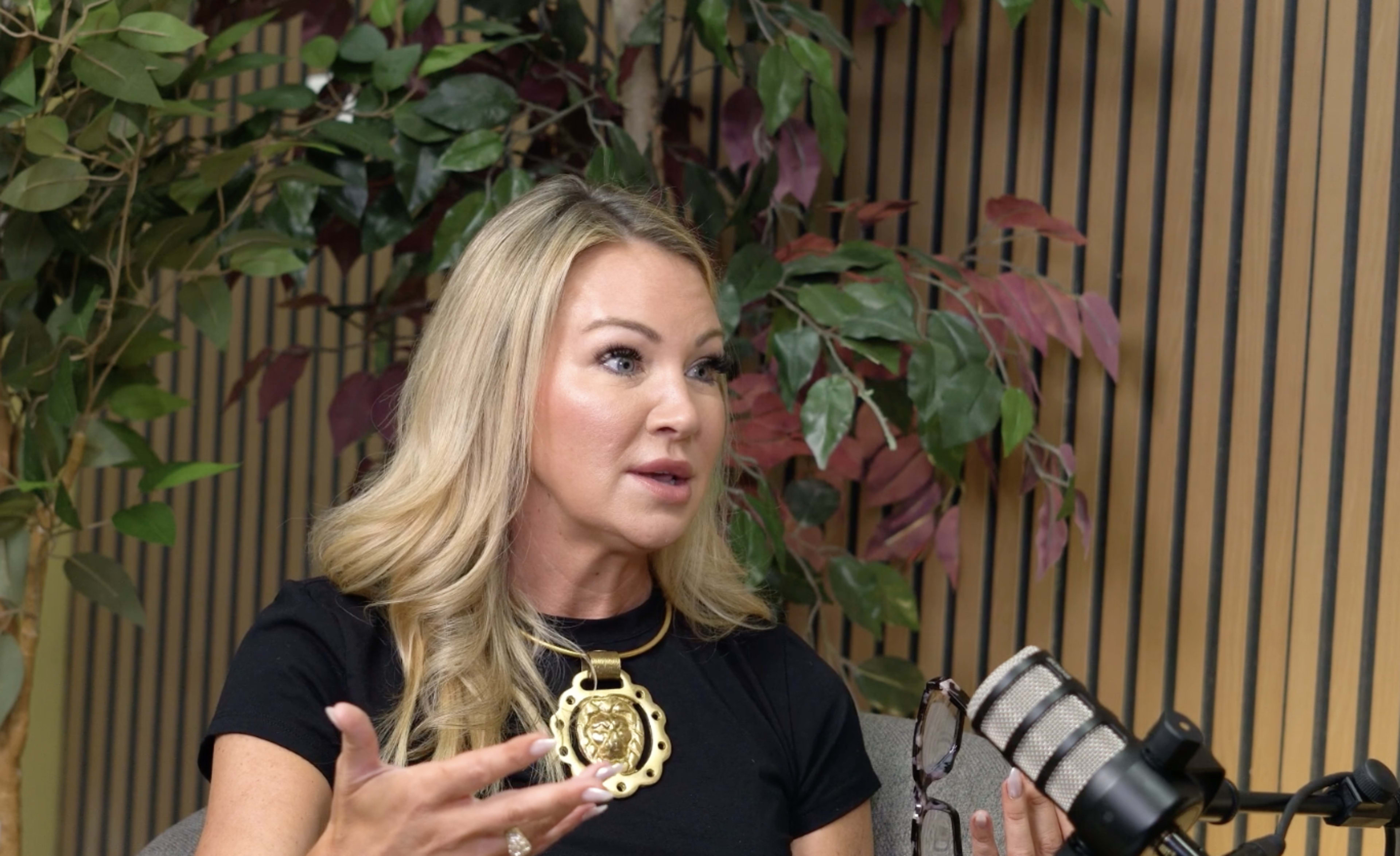 A woman with blonde hair is speaking while sitting in front of a microphone amidst decorative plants.