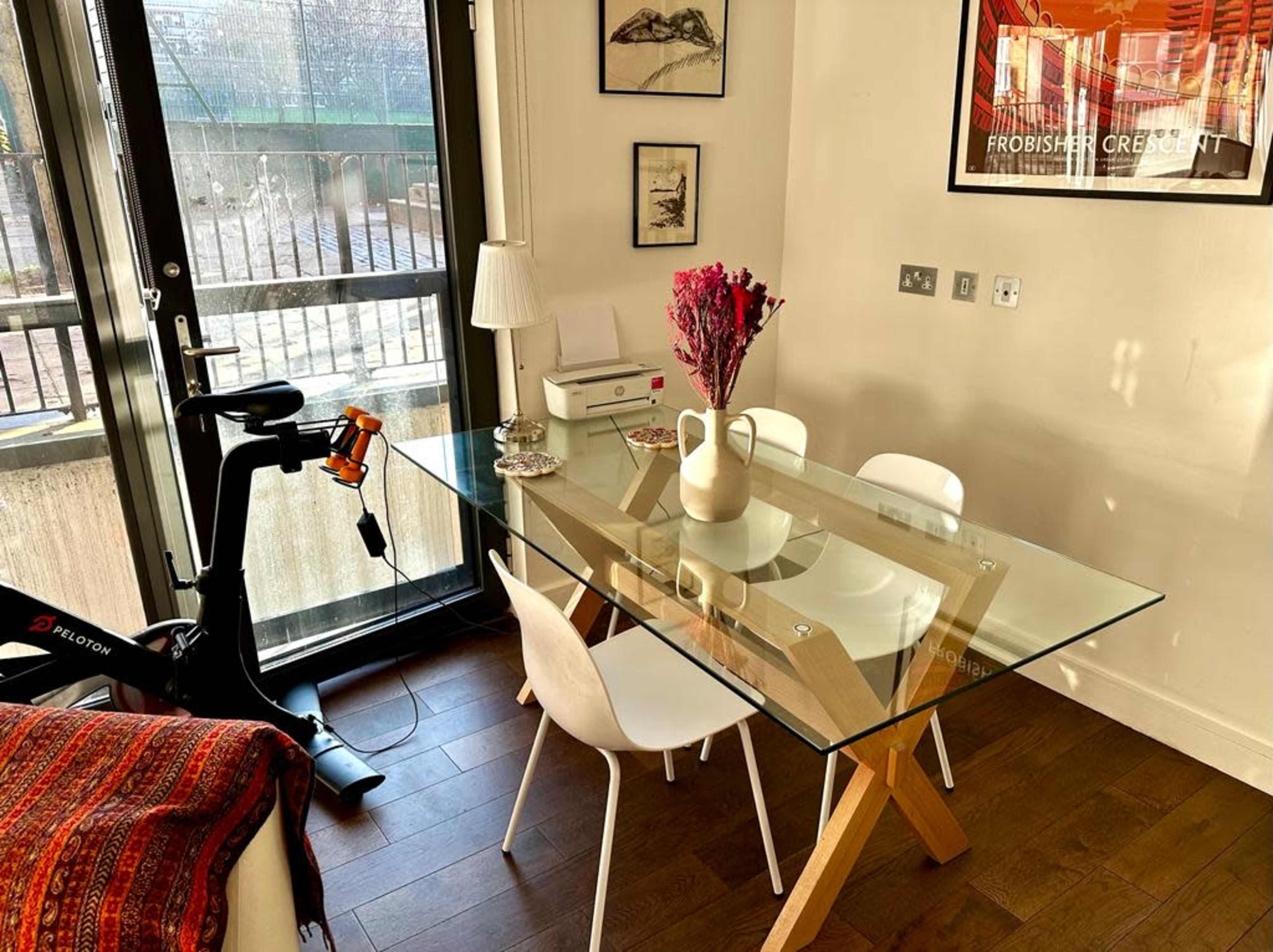 A modern dining area features a glass table with wooden legs surrounded by white chairs, a vase with dried flowers, and an exercise bike nearby.