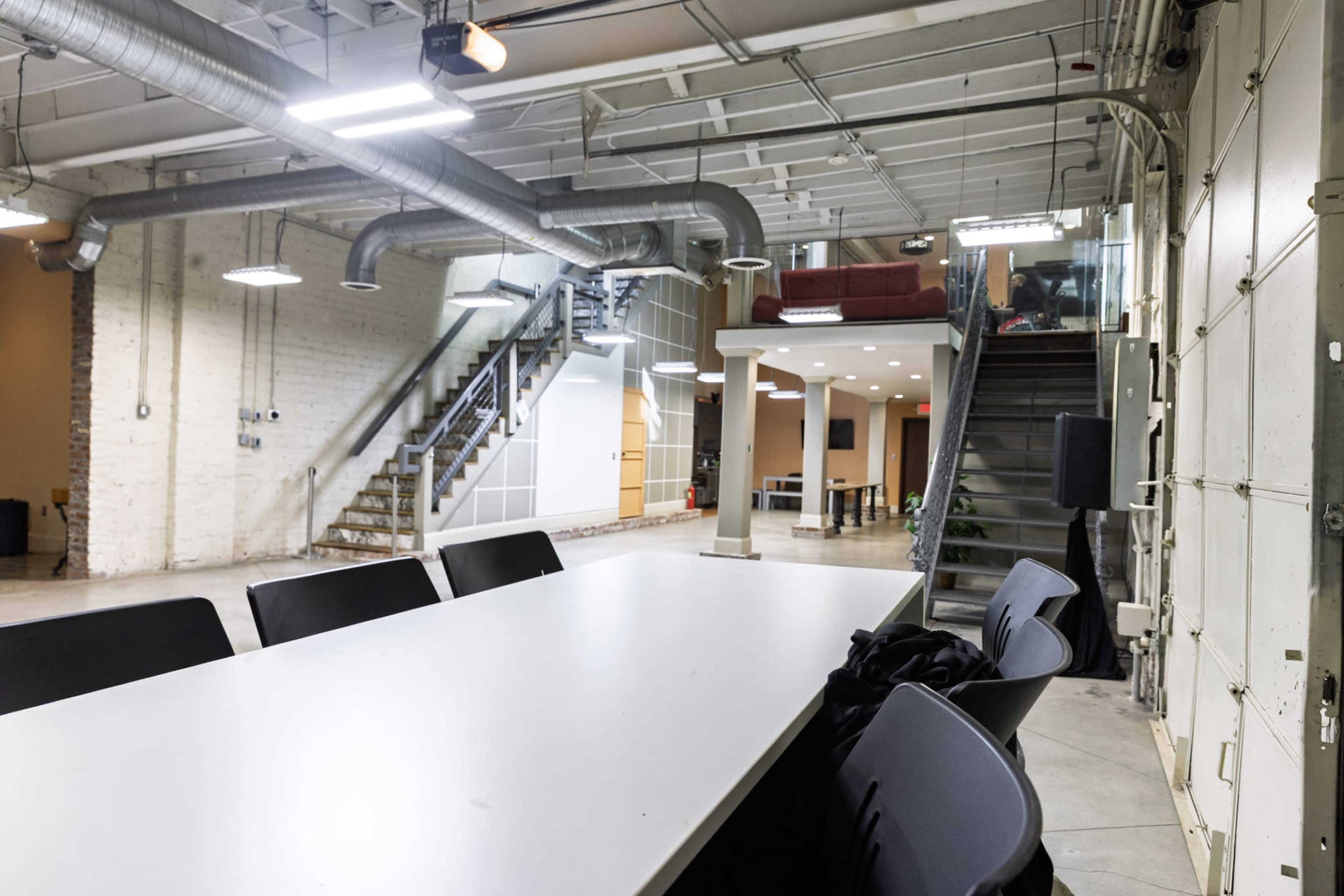 The image shows a large, modern office space featuring a long white table, chairs, and stairs leading to different levels, with exposed ductwork and brick walls.