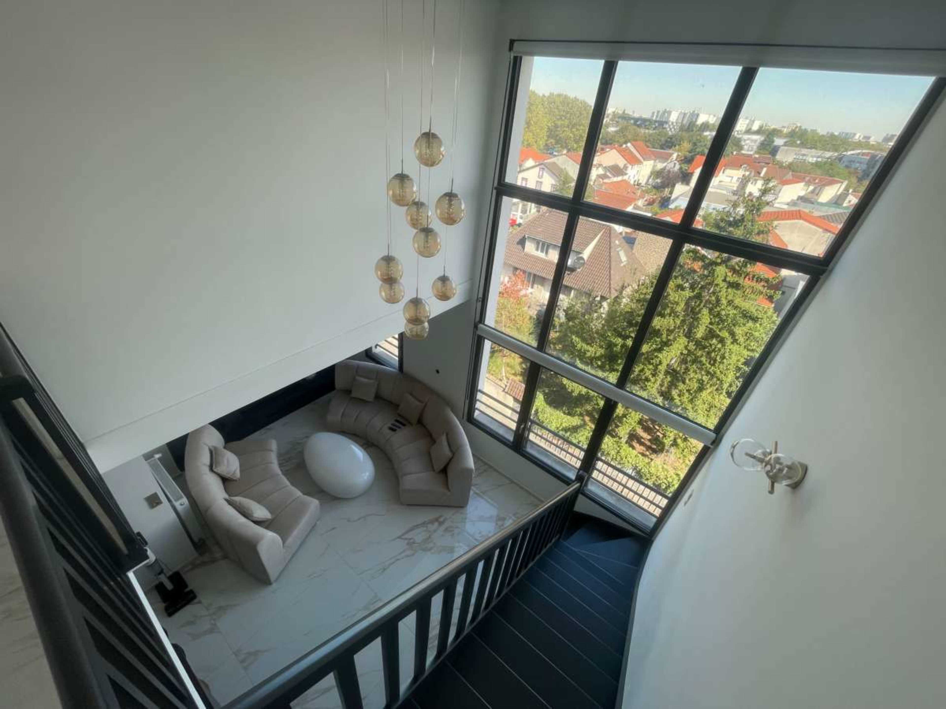 The image shows a modern staircase leading down to a spacious living area with a rounded sofa and a large window overlooking a neighborhood.