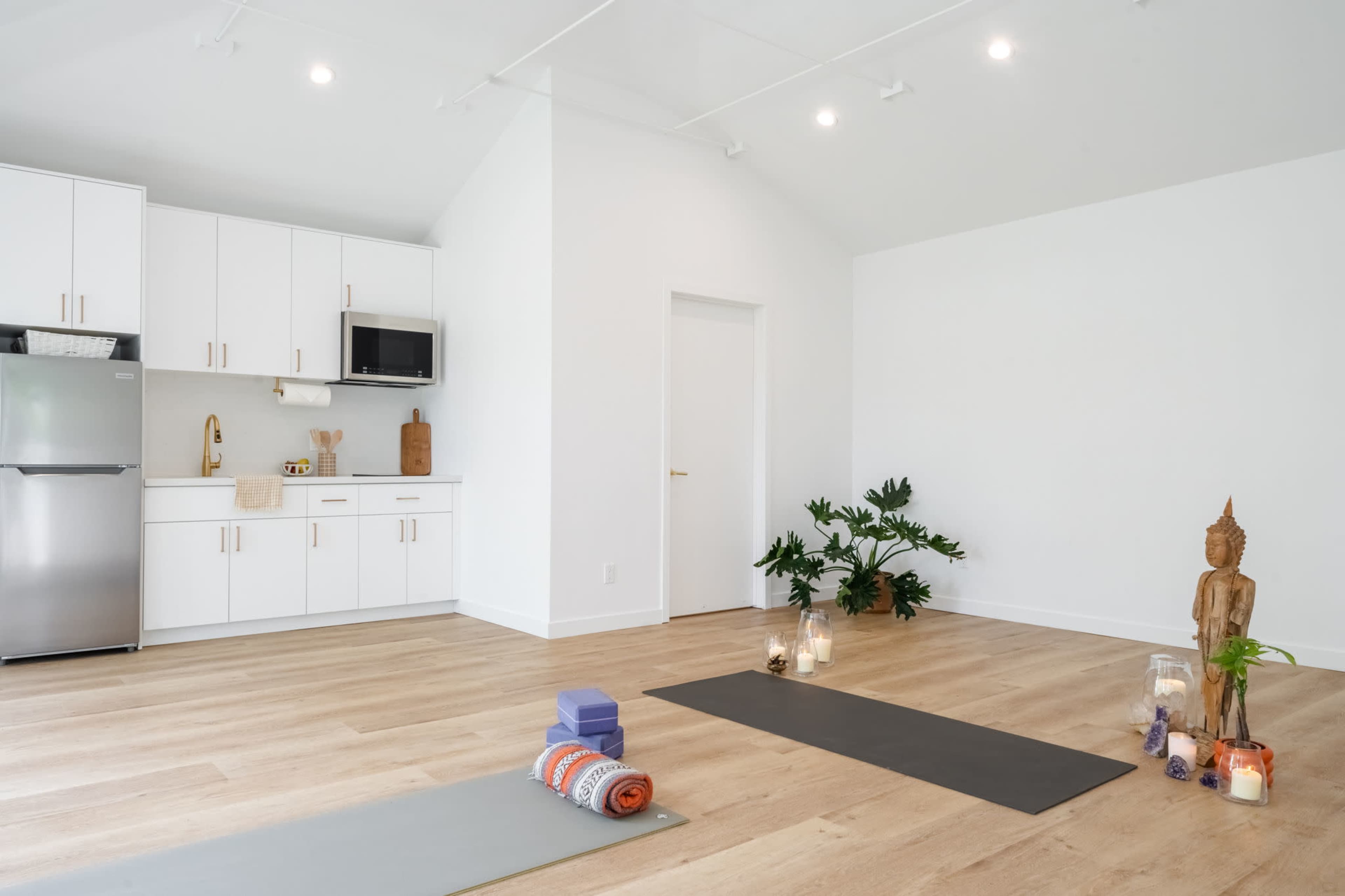 A bright, minimalist yoga studio with two yoga mats, a small kitchen area, and decorative items, including candles and a wooden statue.