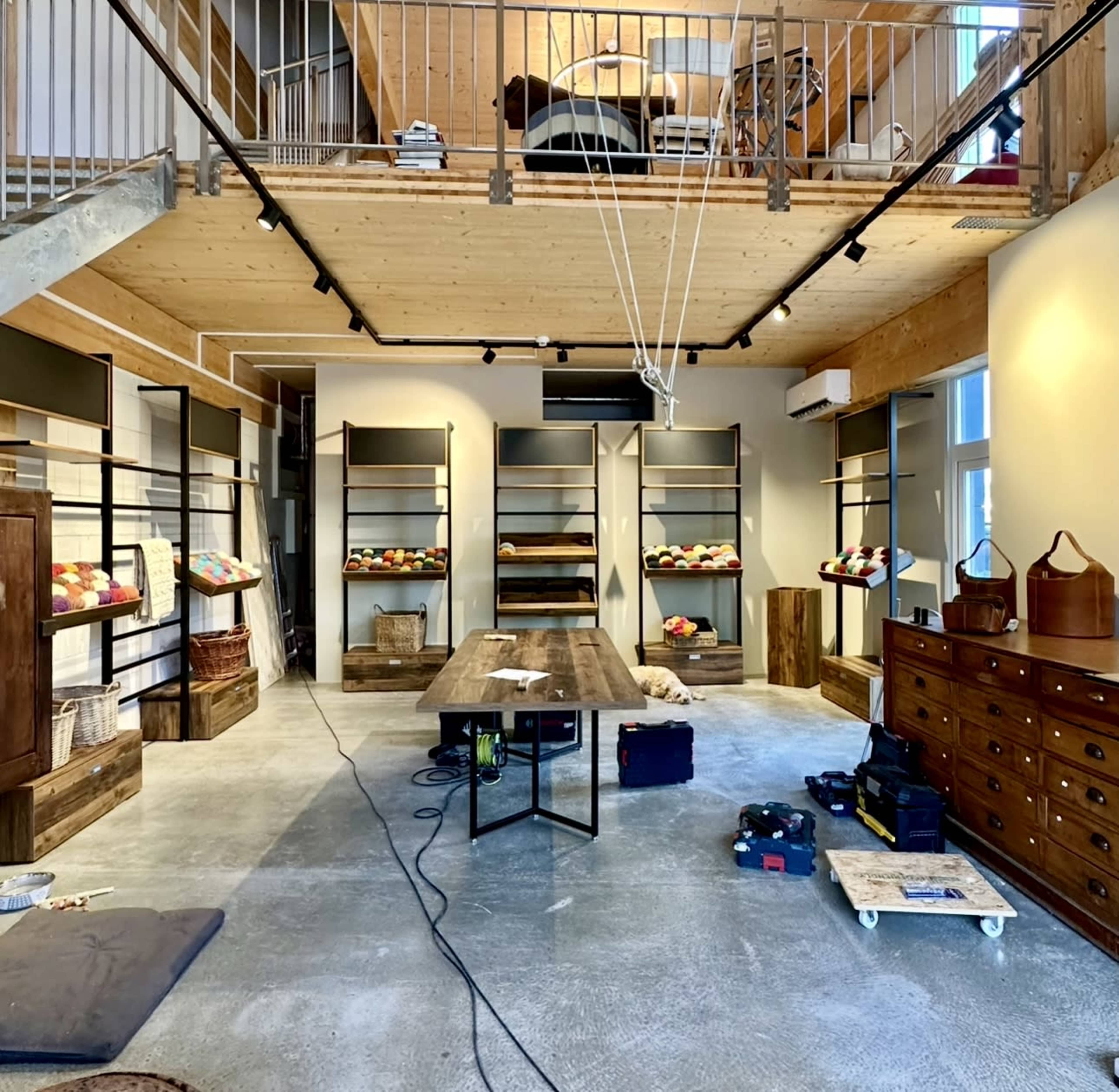 The image shows an interior space with wooden shelves displaying colorful items, a long wooden table in the center, and various tools and equipment scattered on the concrete floor.