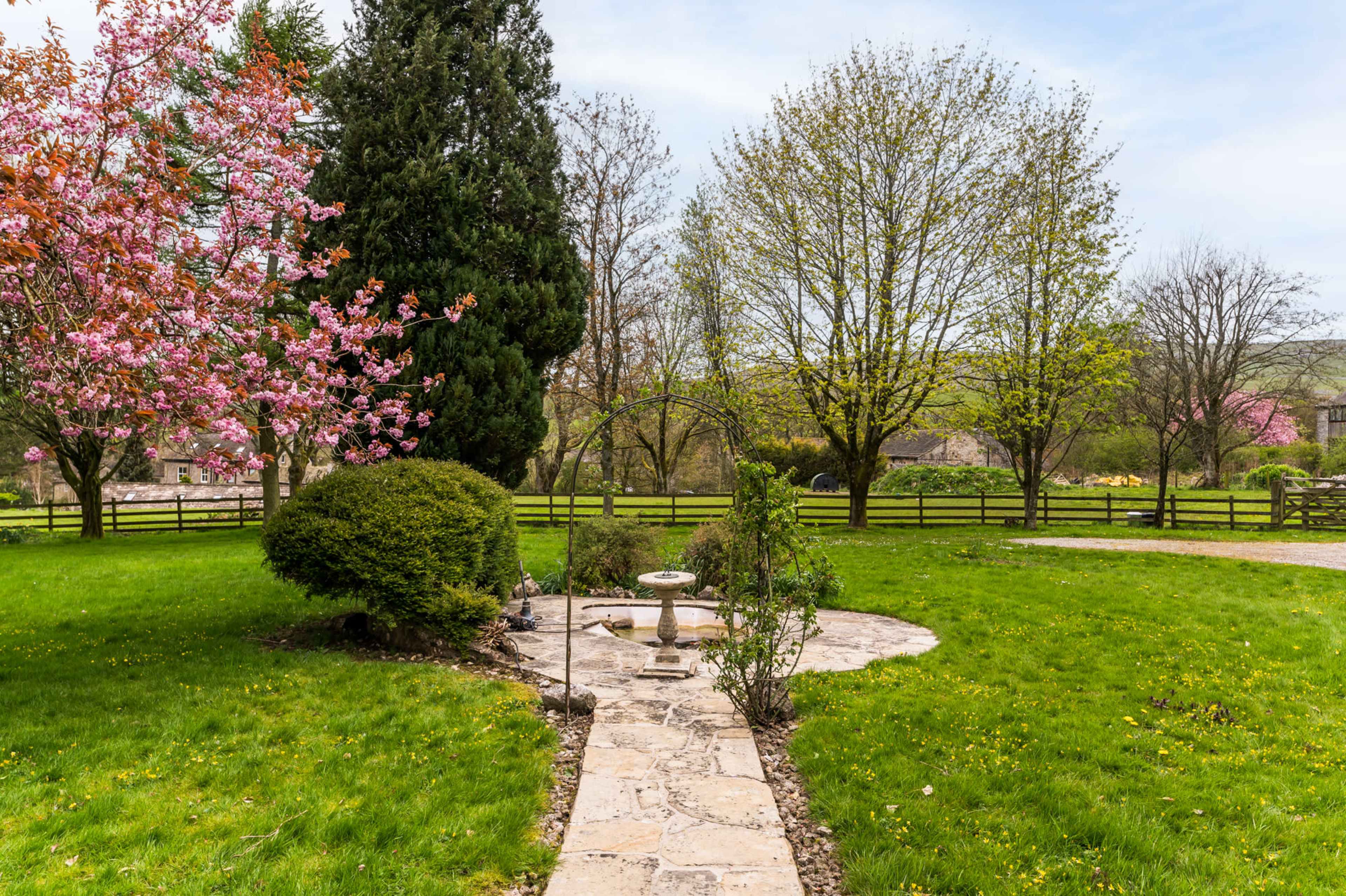 The image shows a landscaped garden with a stone pathway leading to a birdbath, surrounded by greenery and trees, including a pink flowering tree.