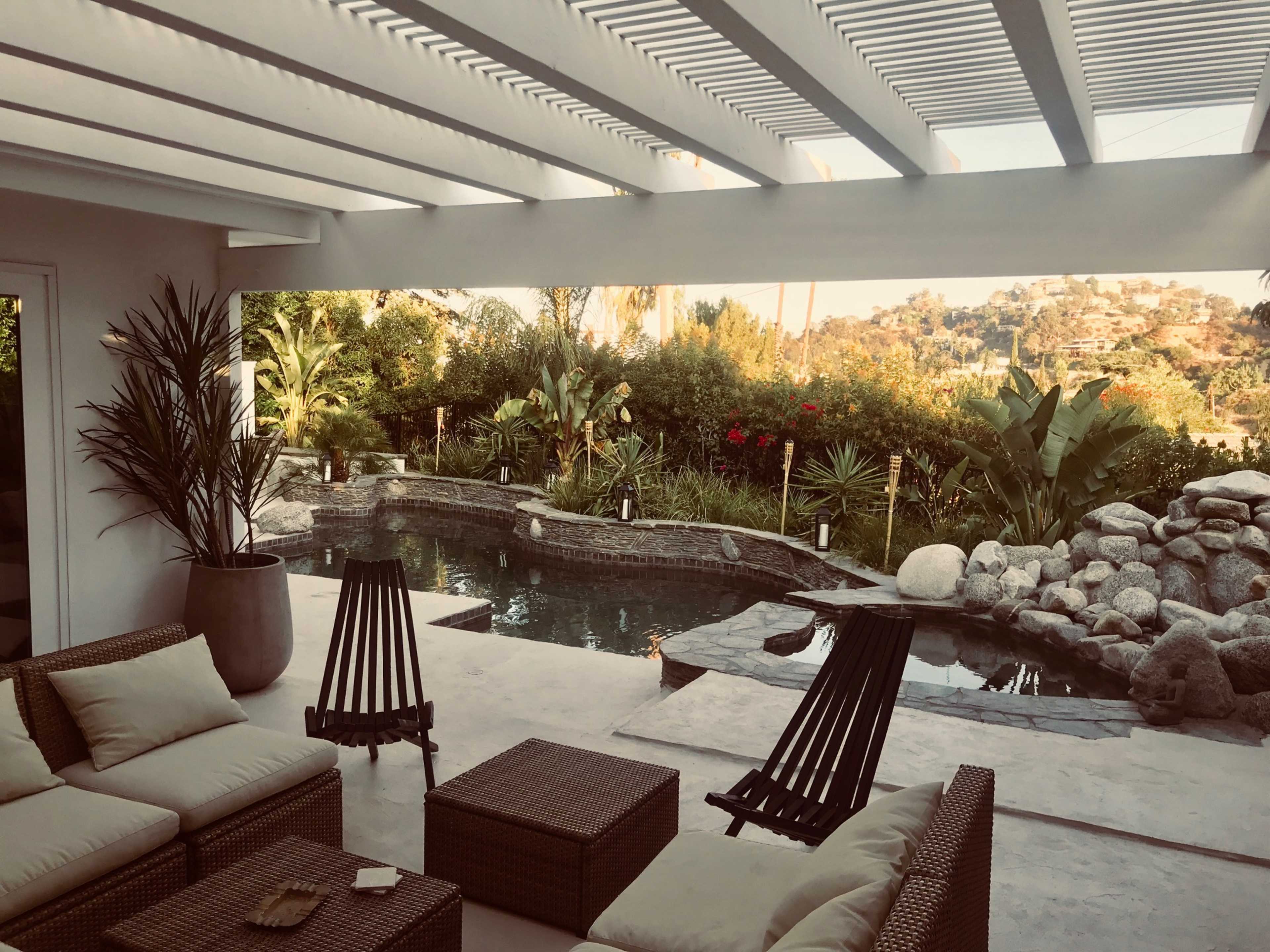 A covered patio with lounge chairs surrounds a natural-looking pool, bordered by tropical plants and rocks.