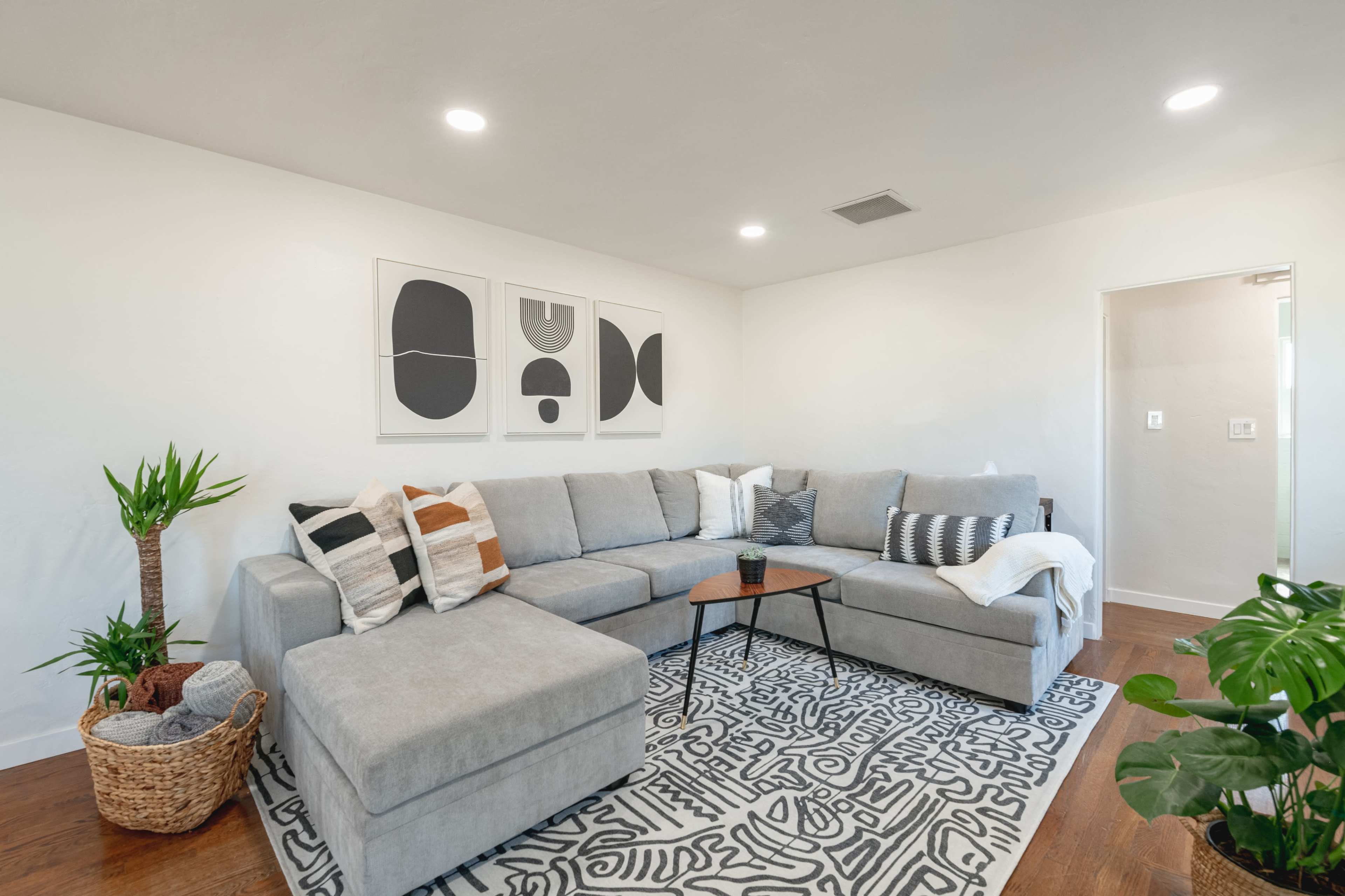 A gray sectional couch with a variety of decorative pillows is arranged on a patterned rug in a well-lit living room featuring modern artwork on the walls and indoor plants nearby.