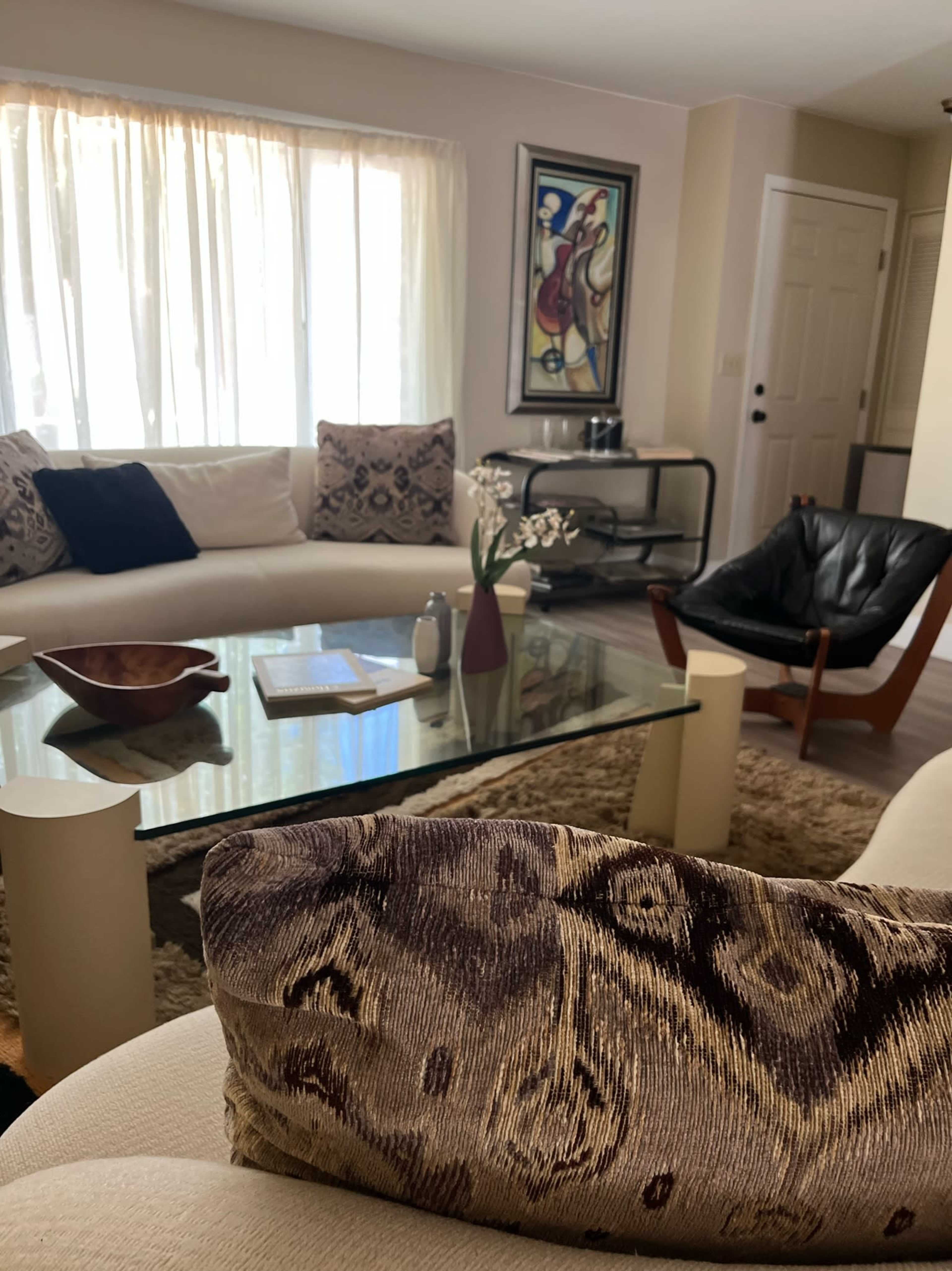 A light-filled living room features a sectional sofa, a glass coffee table, and various decorative items.