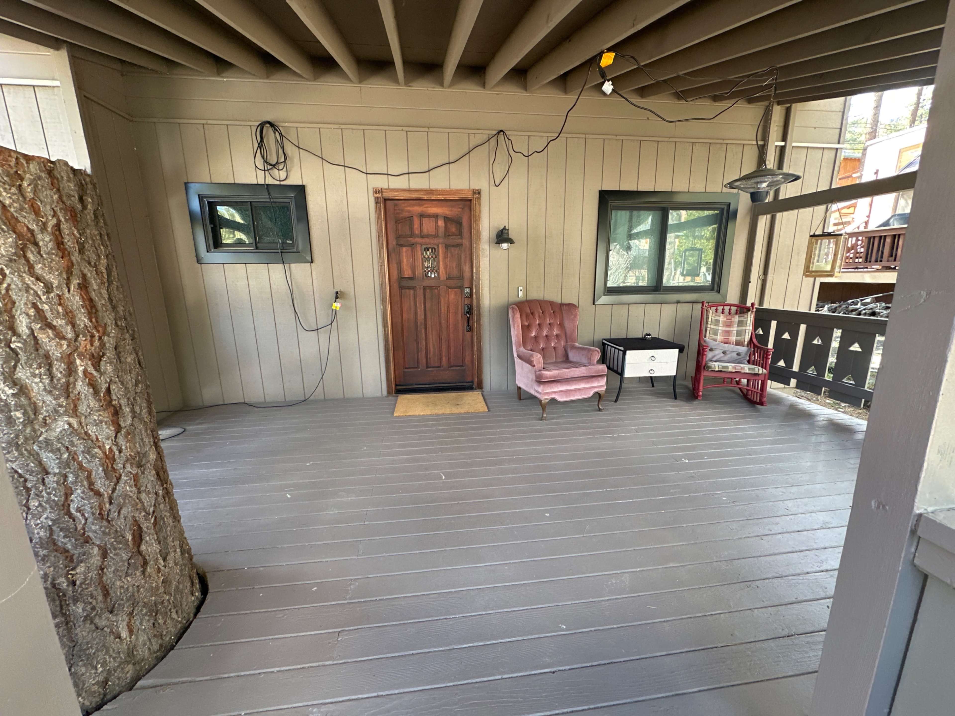 A covered porch featuring a wooden door, a pink armchair, a small table, and a tree column on one side.
