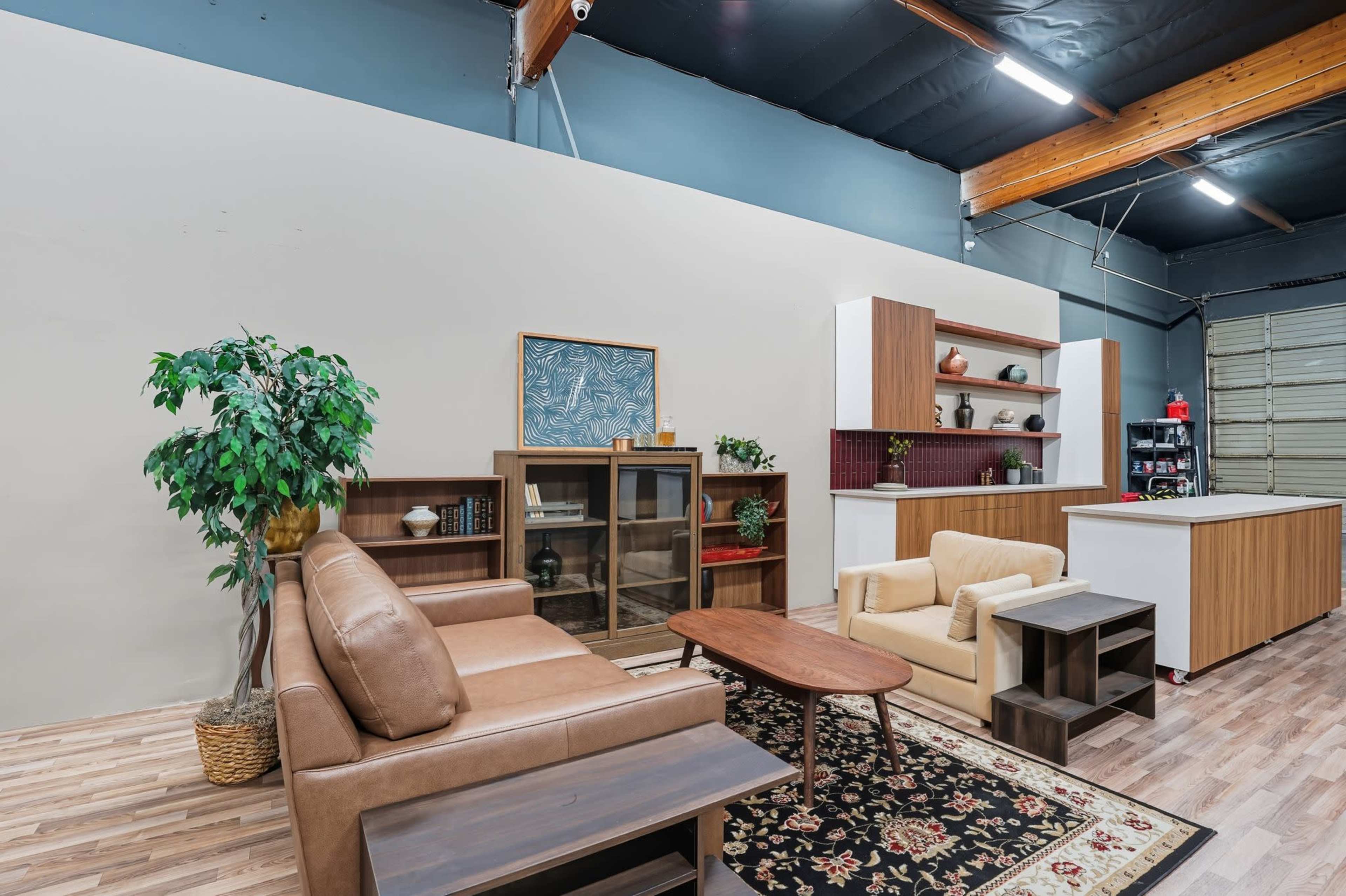 The image shows a modern interior space featuring a brown leather sofa, a beige armchair, a wooden coffee table, and decorative shelving units against a light-colored wall.
