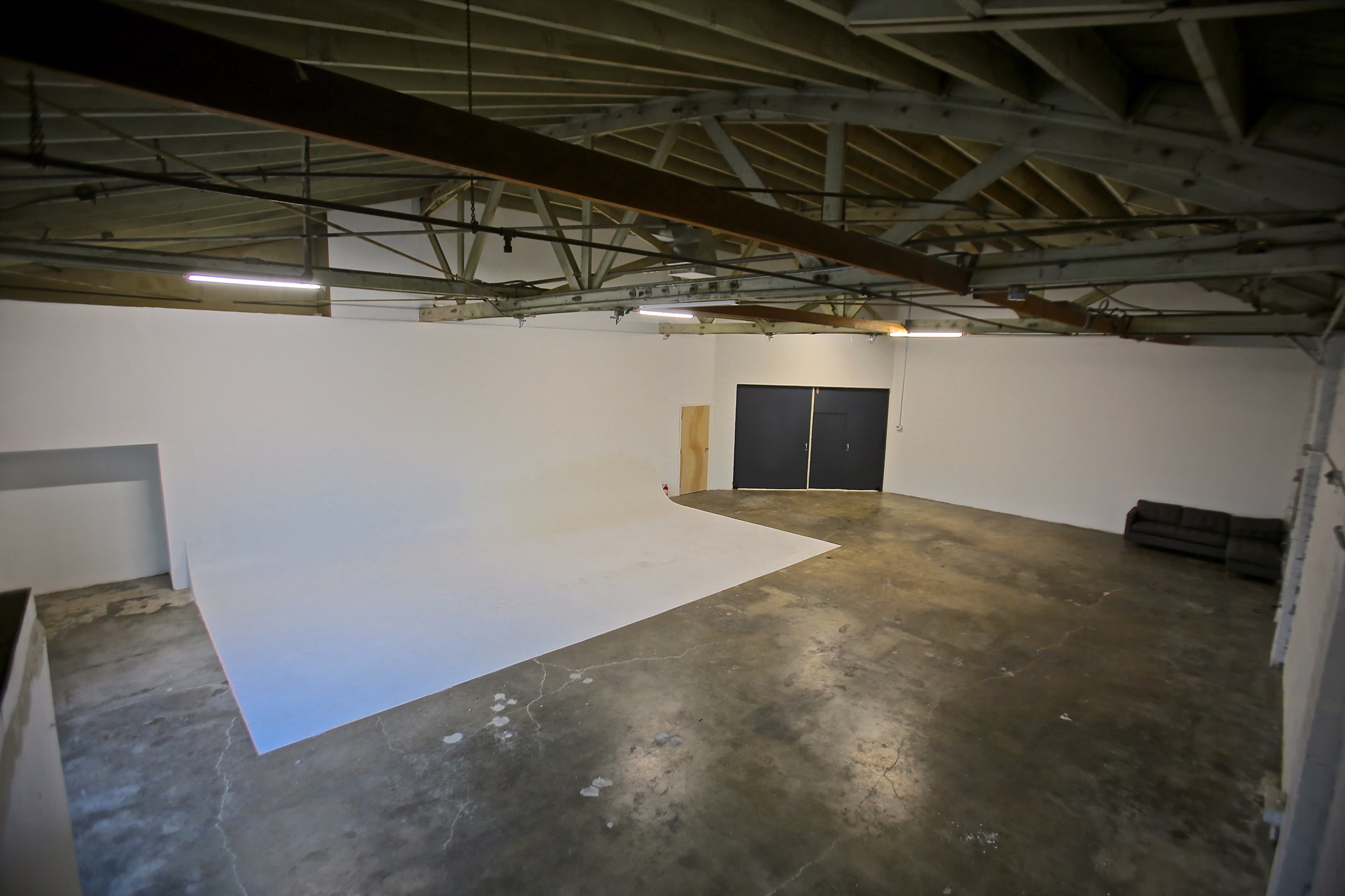 A large, empty studio space with a white backdrop, concrete flooring, exposed beams, and a black couch against one wall.