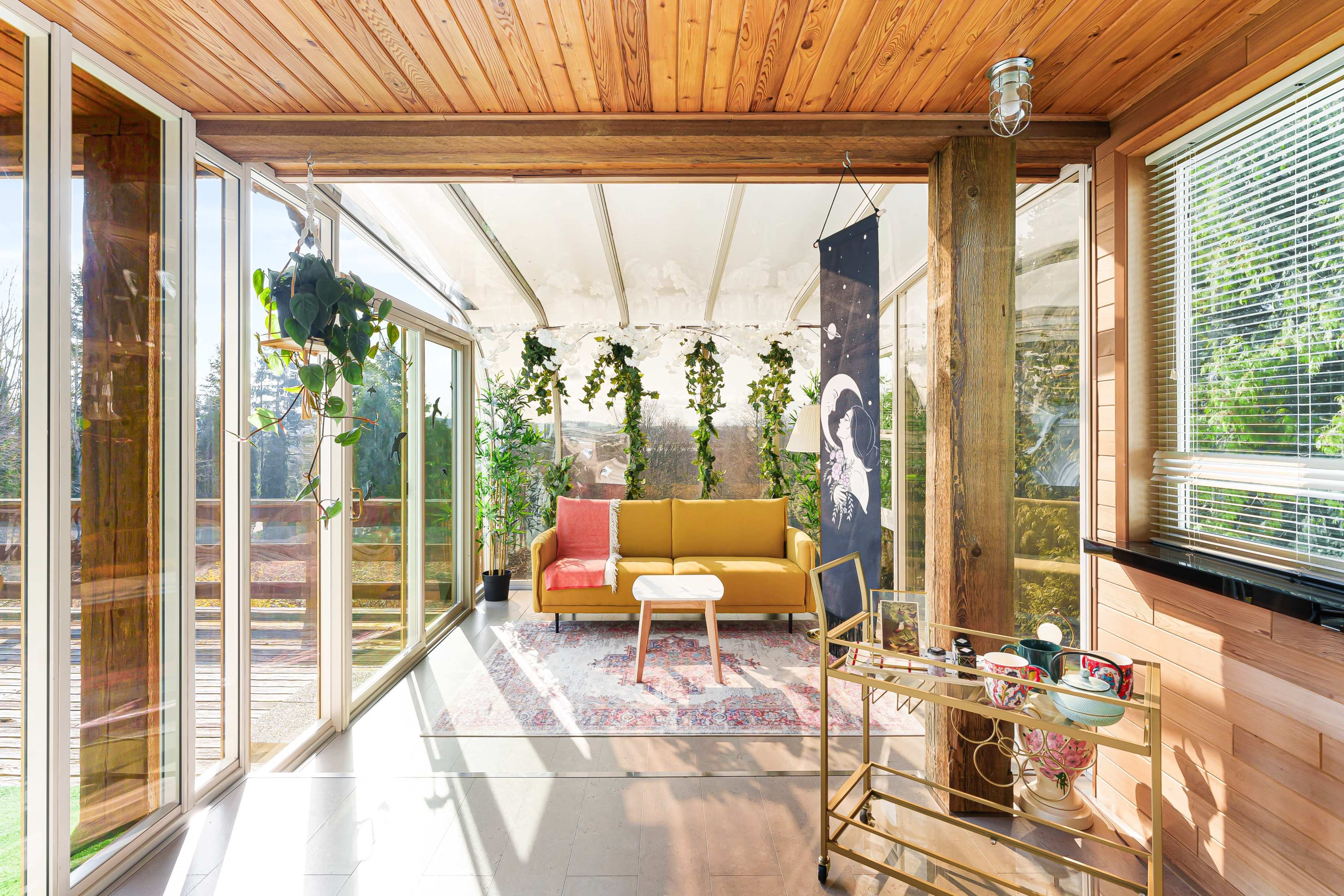 A sunlit sunroom with a yellow sofa, a small coffee table, and plants decorating the space, alongside a cart holding various items.