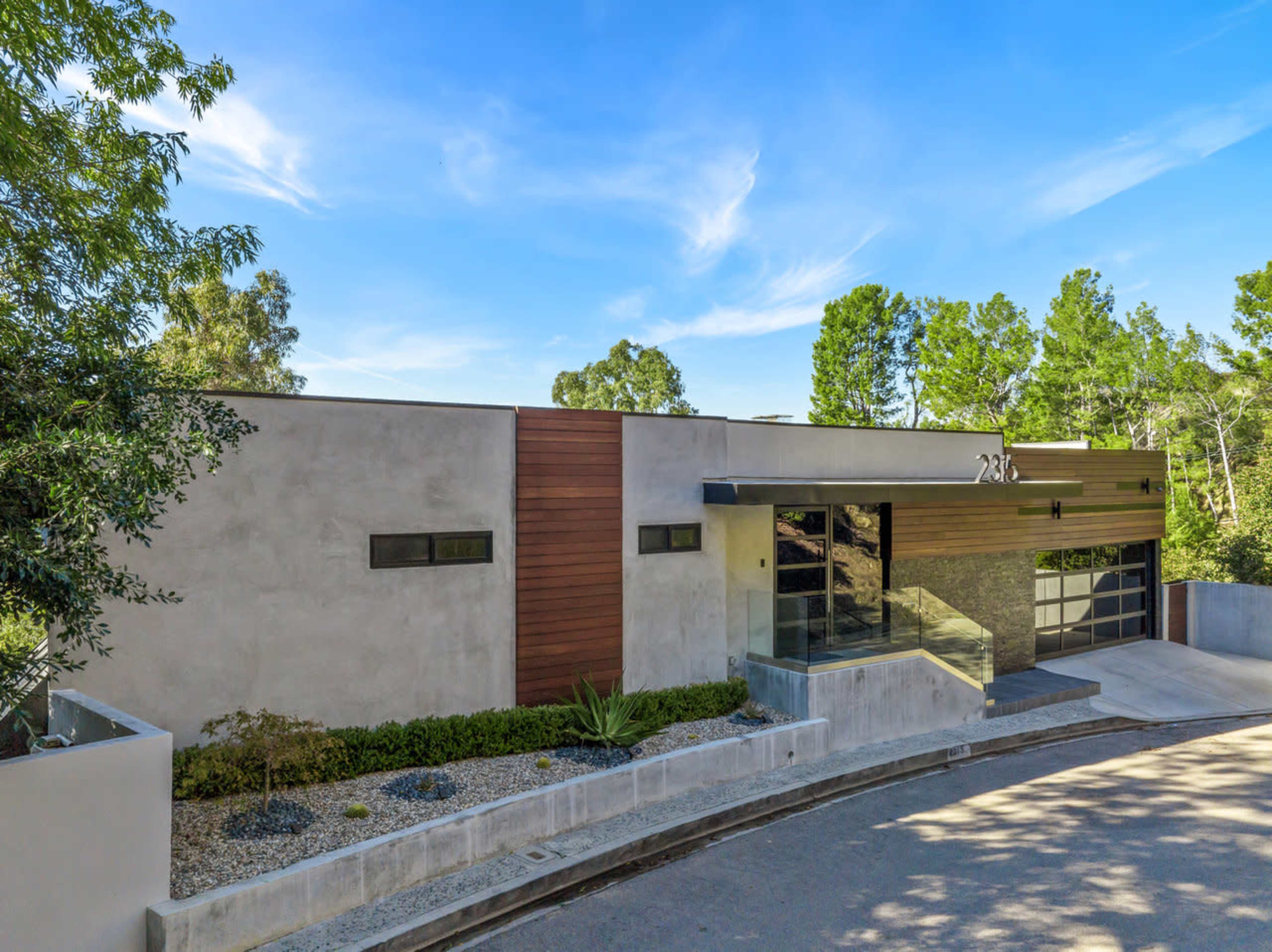 A modern single-story house with a mixture of textured concrete and wood accents is situated on a curved driveway surrounded by greenery.