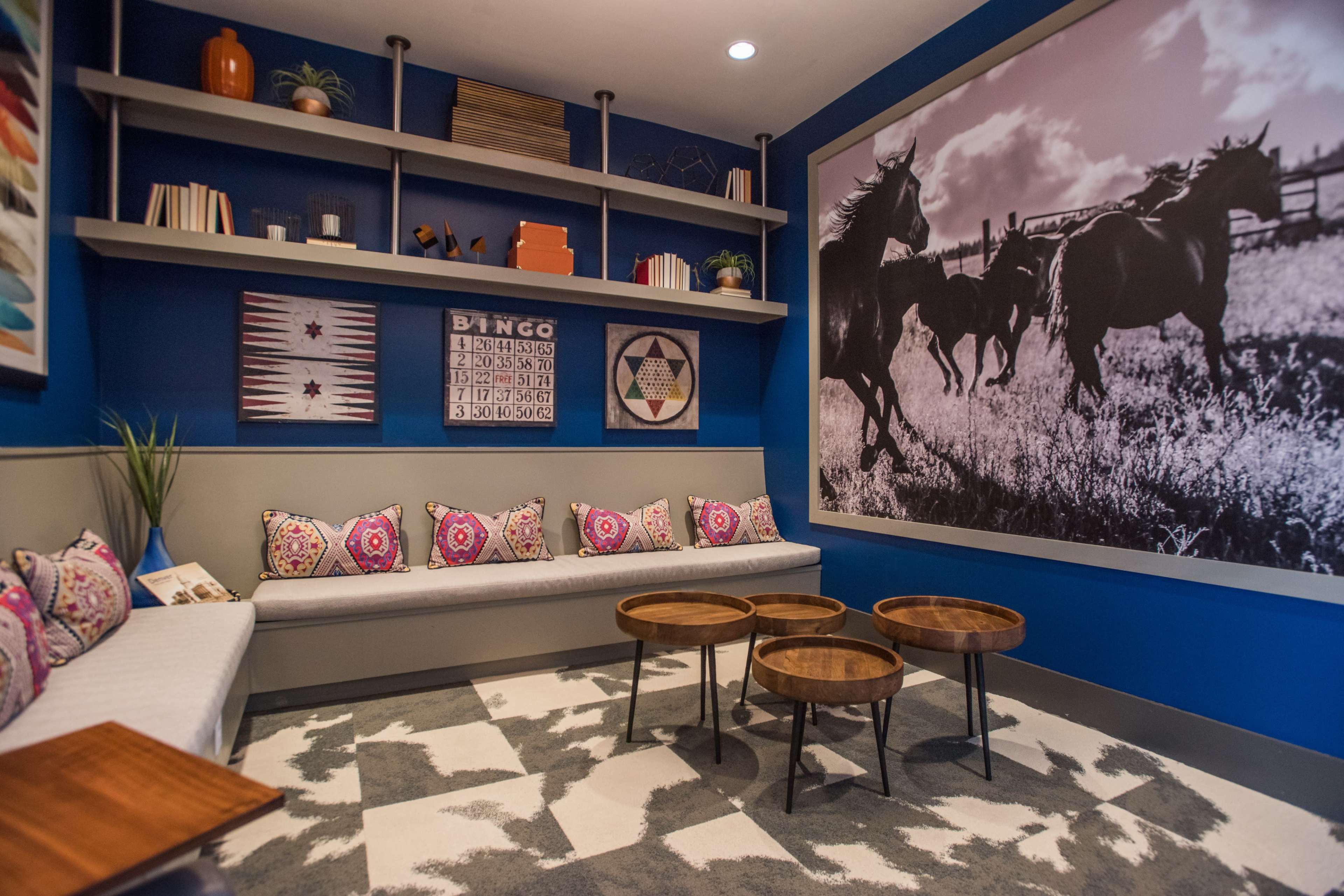 A cozy lounge area with a blue accent wall, a large black-and-white horse photograph, shelves with decorative items, and a seating area featuring colorful cushions and wooden coffee tables.