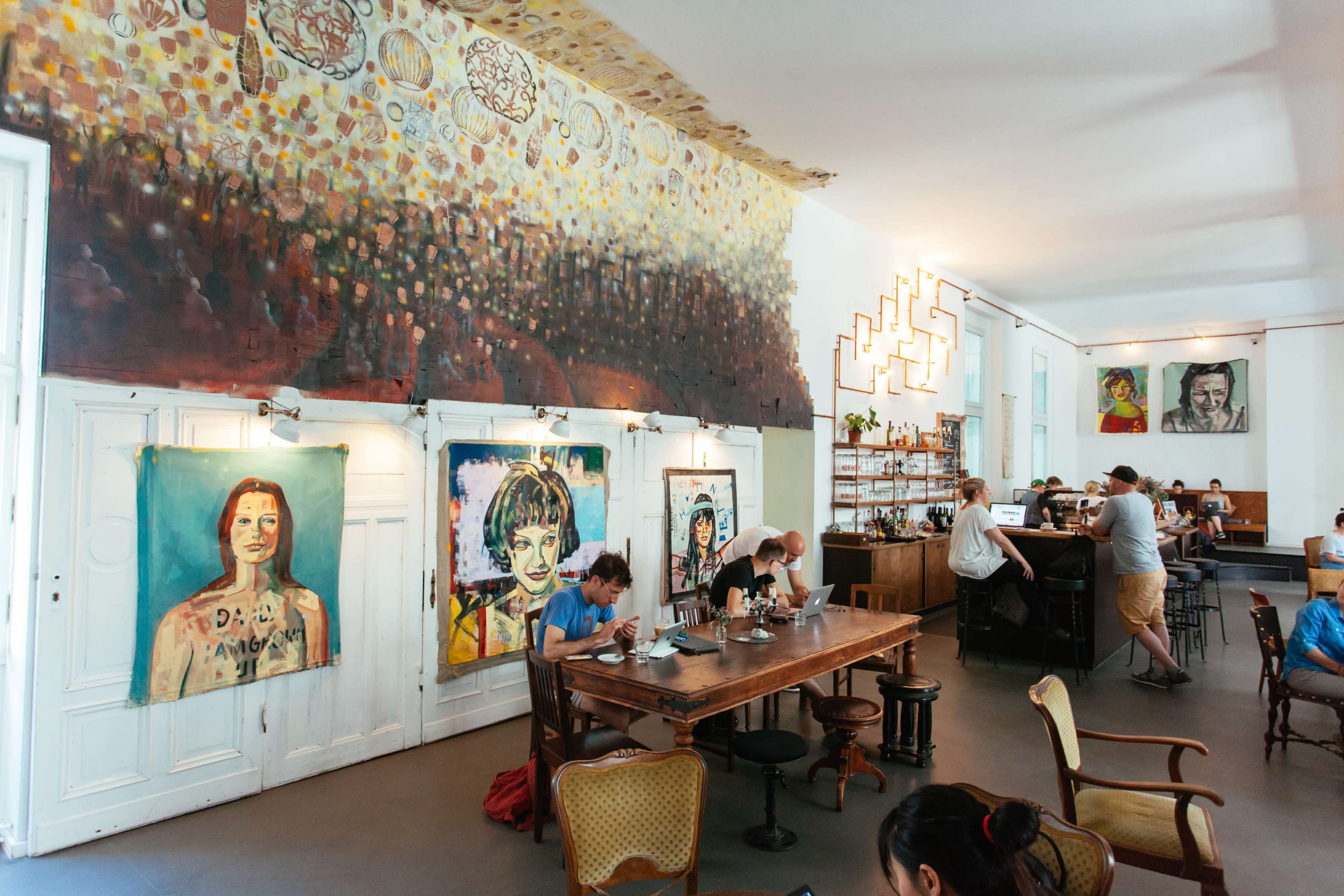 A café interior featuring large colorful paintings on the walls, a wooden dining table, and customers sitting at tables and the bar.