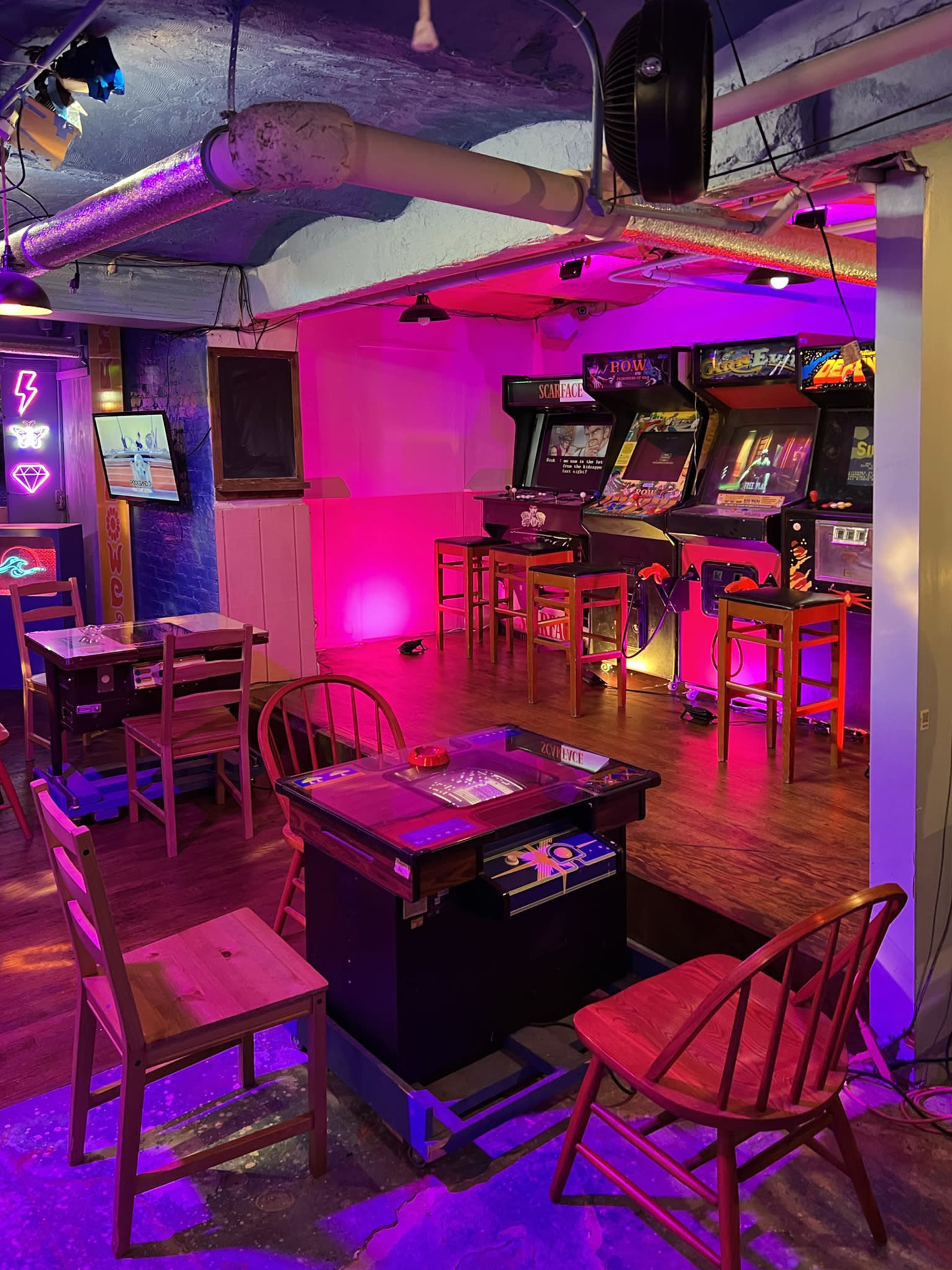 A retro arcade with multiple video game machines and tables, illuminated by colorful lights.