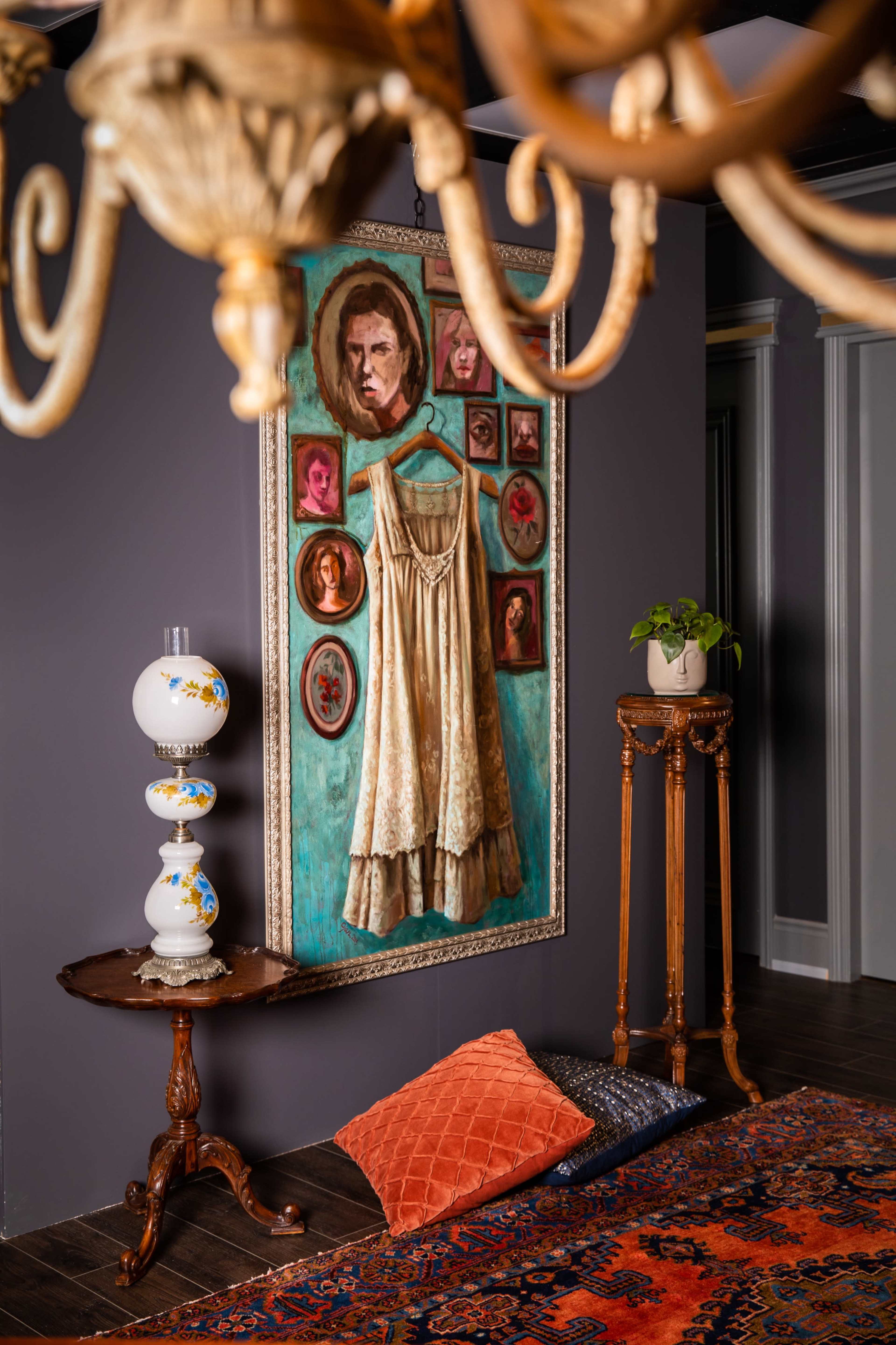 The scene features a wall adorned with a large portrait and smaller framed images, a vintage lamp on a table, and a patterned rug with decorative cushions on the floor.