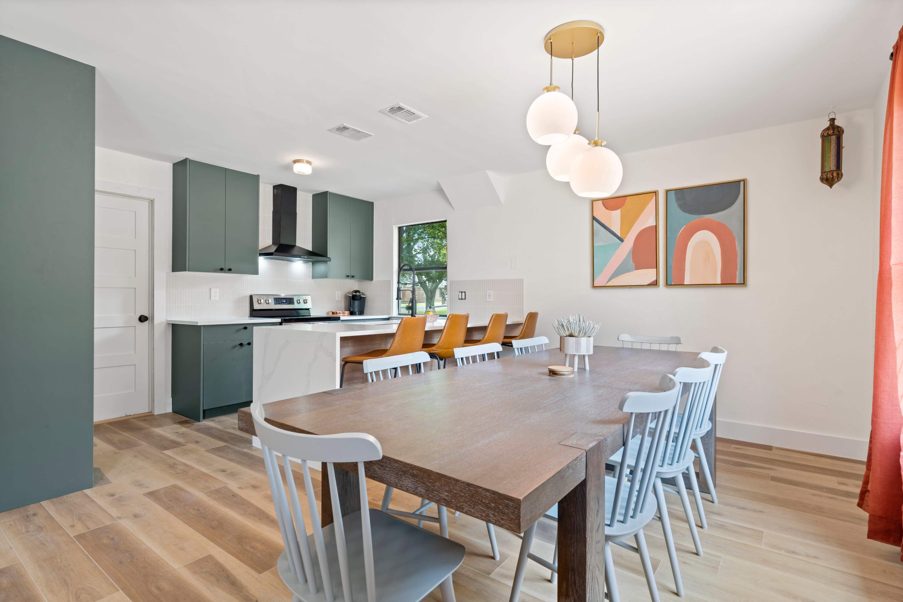 A large dining table with light gray chairs is positioned in a modern kitchen featuring dark green cabinets, a white countertop, and art on the walls.