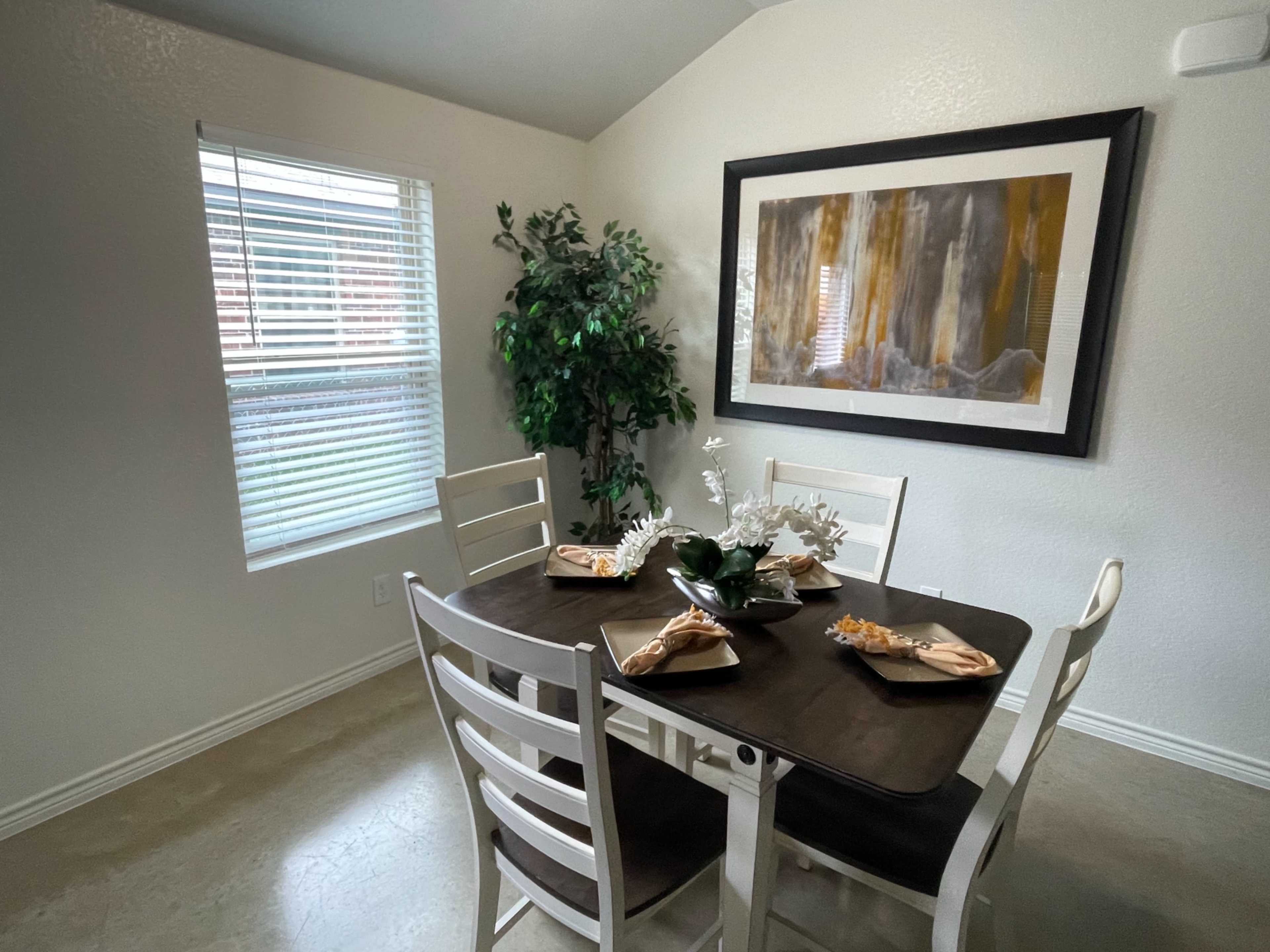 A dining area with a brown table set for four, adorned with decorative plates and a centerpiece, alongside a window and a potted plant.