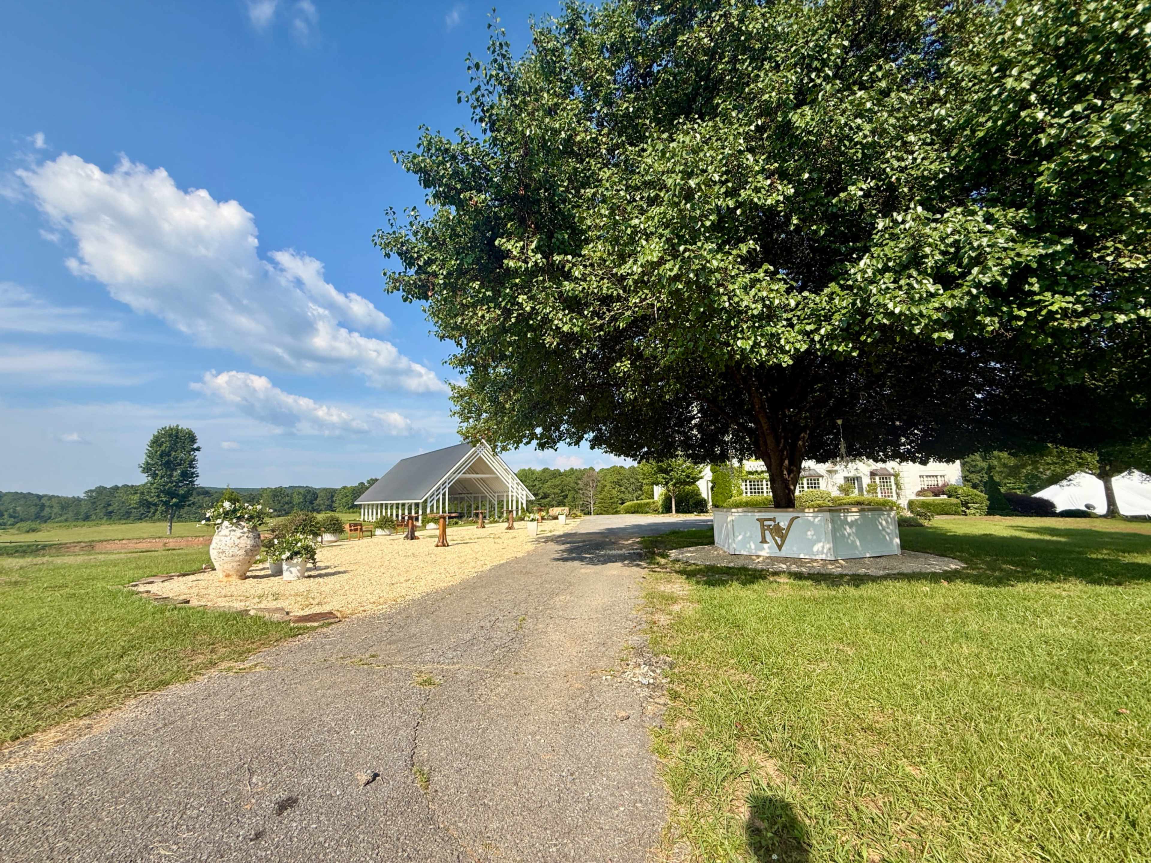 A gravel path leads to a white tent structure near a large tree, with a house visible in the background under a clear blue sky.