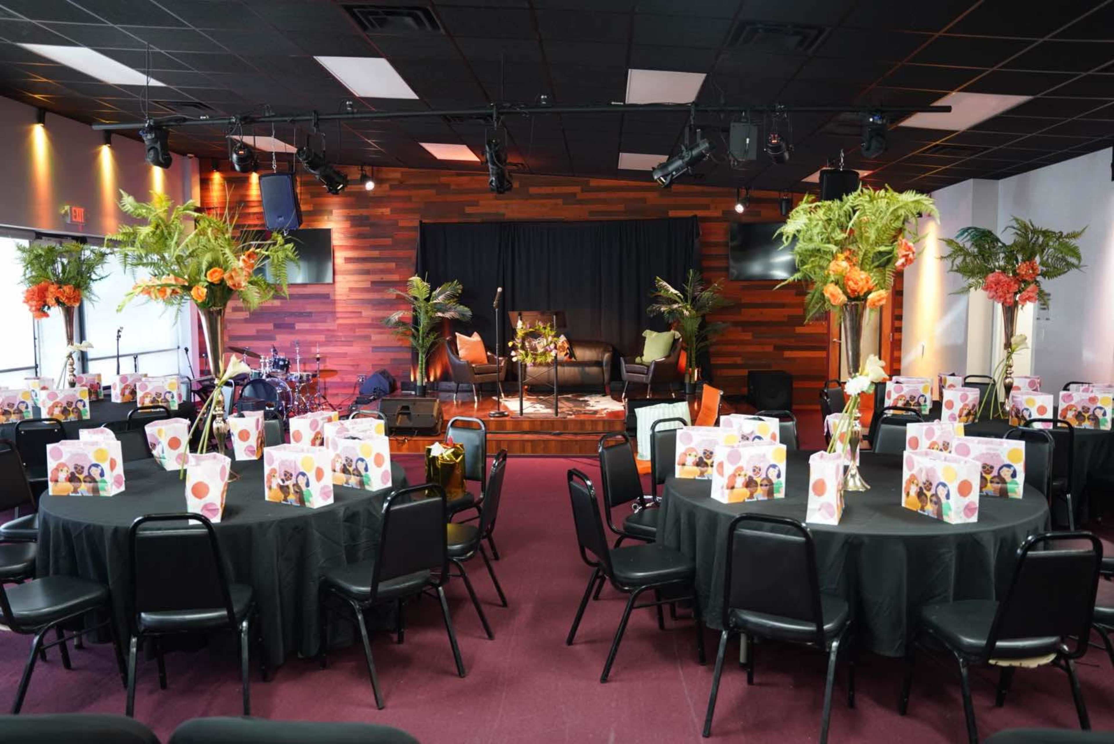 The image shows a banquet hall set up for an event, with several round tables covered in black tablecloths and decorated with floral arrangements, while the stage features a backdrop and seating.