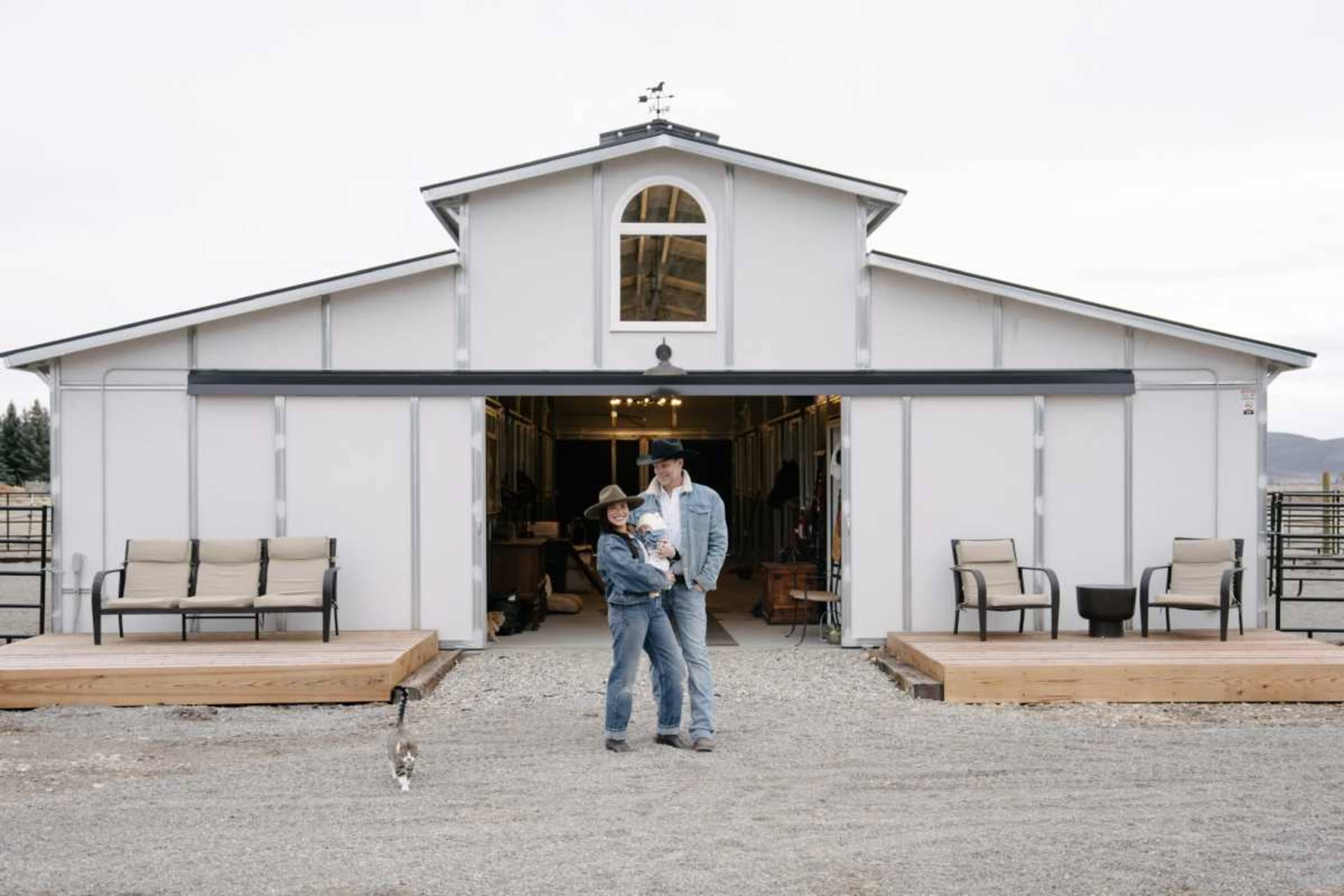 A person holding a child stands in front of a large barn-like structure, while a cat walks nearby on a gravel surface.