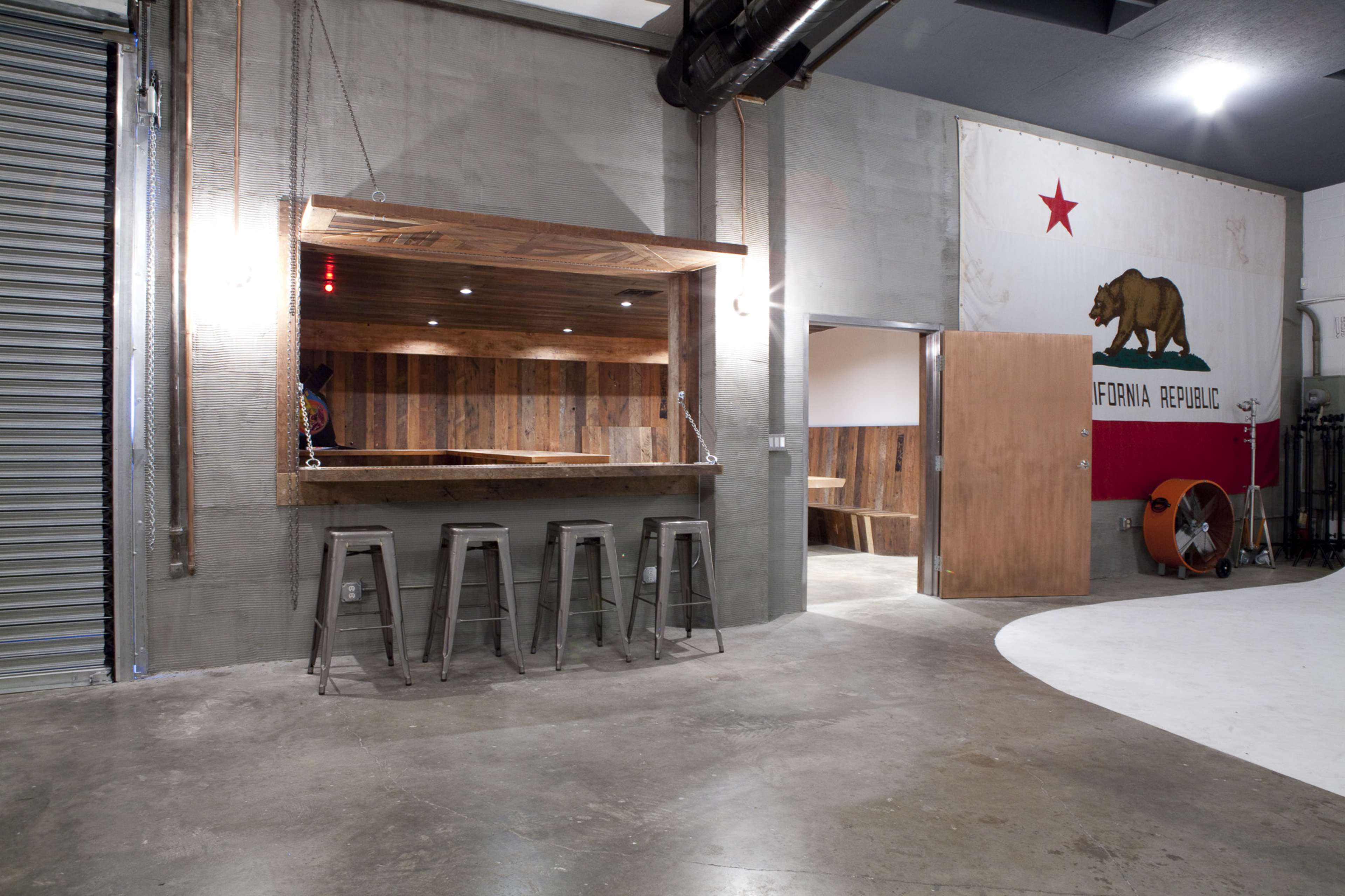 A modern industrial space featuring a wooden bar with stools, a rolling metal door, and a wall adorned with a California flag and bear emblem.