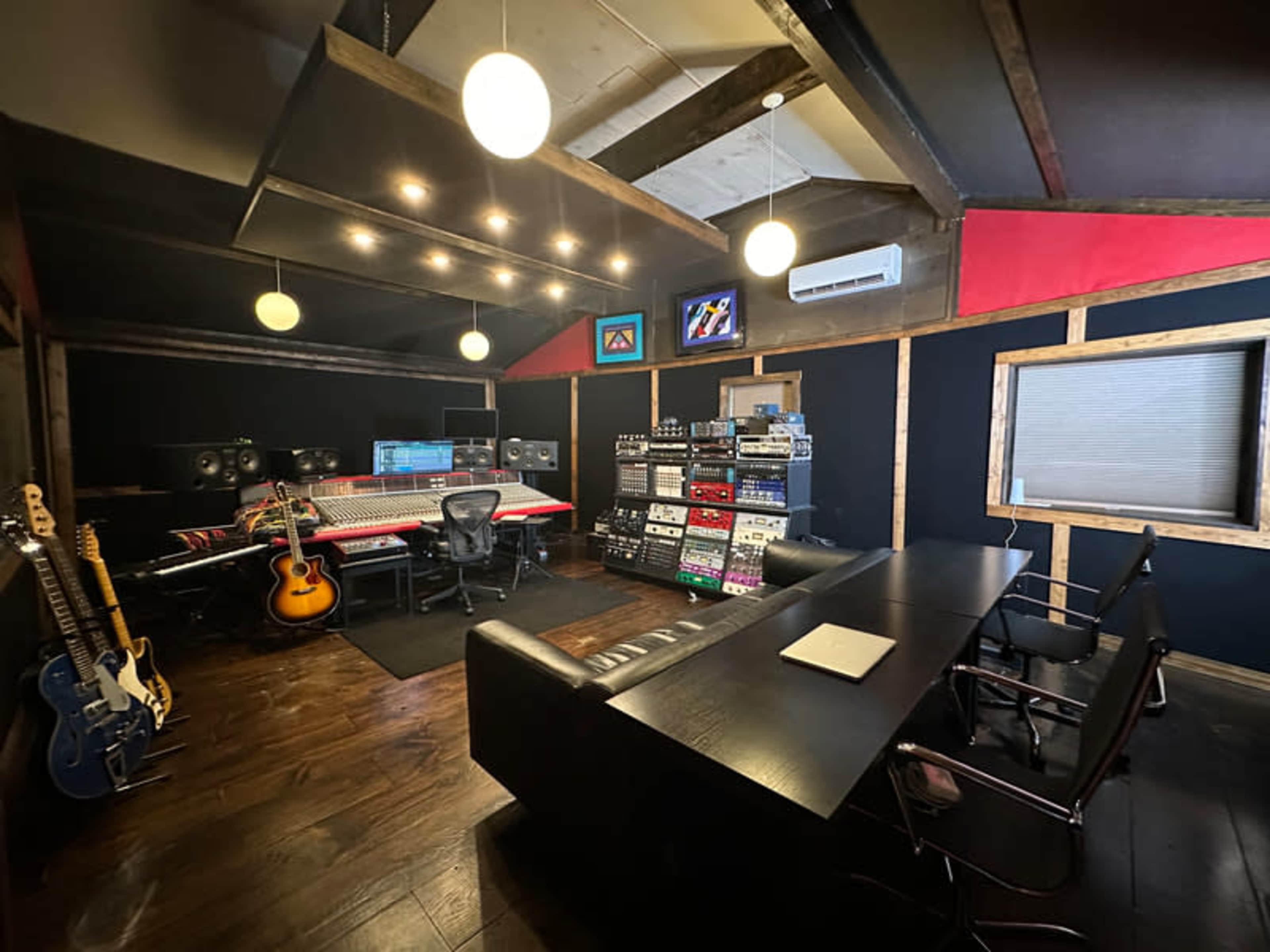 A music production studio featuring a central mixing console, audio equipment racks, several guitars, and a conference table with chairs.