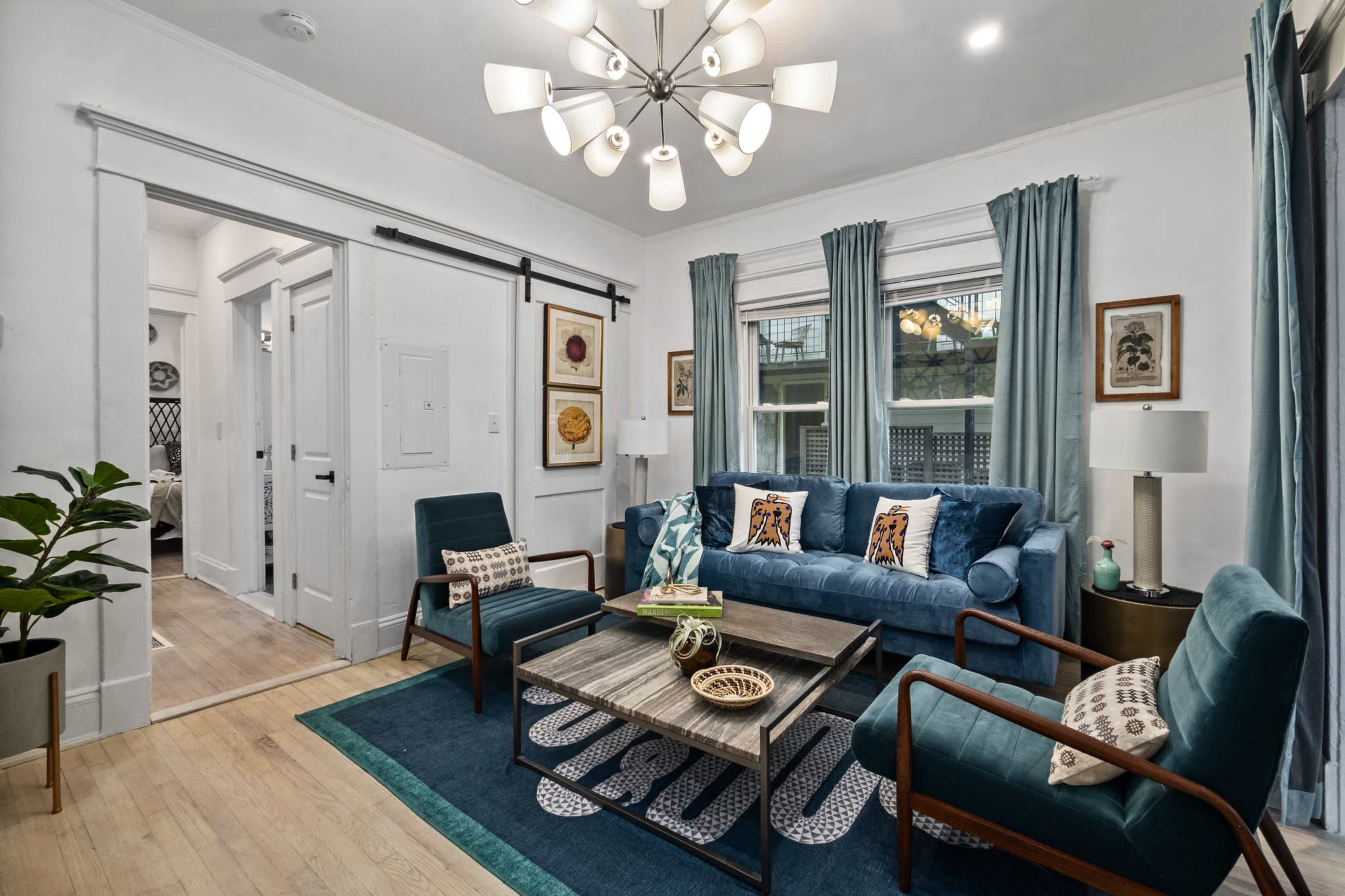 The image shows a well-decorated living room featuring a blue sofa, two green chairs, a wooden coffee table, and large windows with sheer curtains.