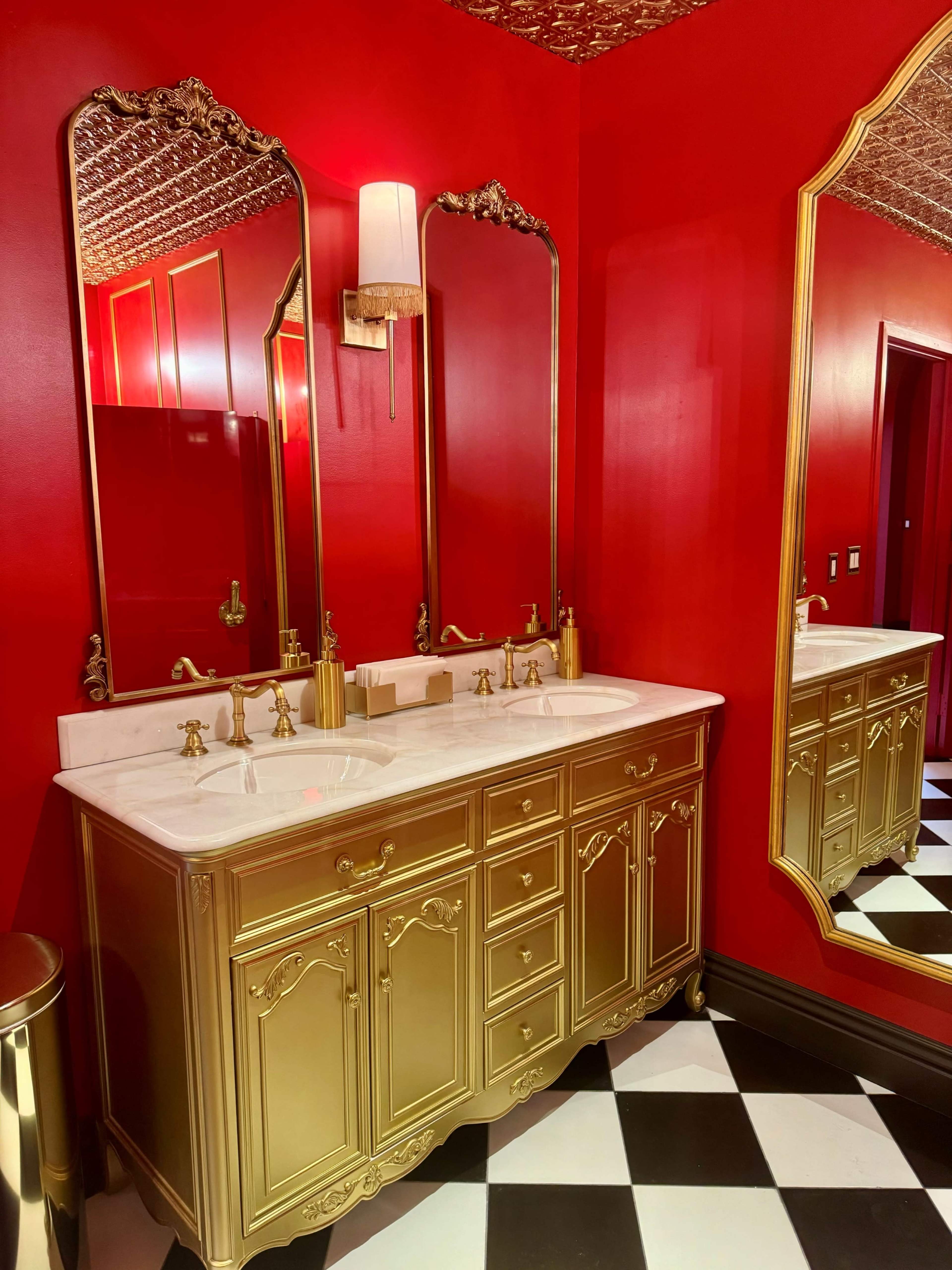 The bathroom features a gold vanity with a marble countertop, dual sinks, and ornate mirrors against a vivid red wall and a black-and-white checkered floor.