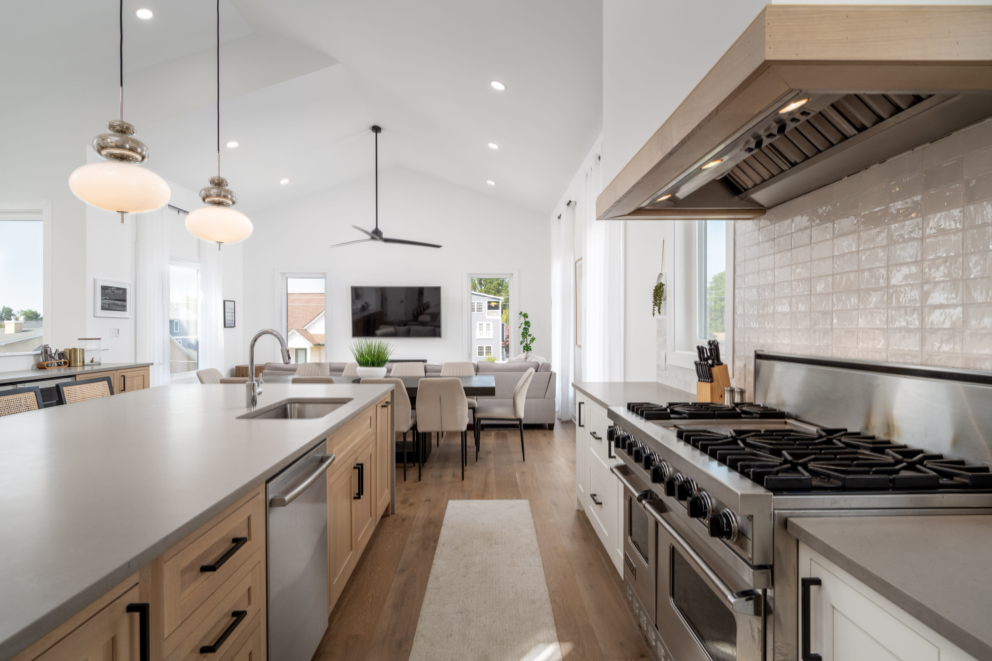 A modern kitchen with wooden cabinets, a long countertop, and a professional-grade stove opens up to a dining area and living space with large windows.