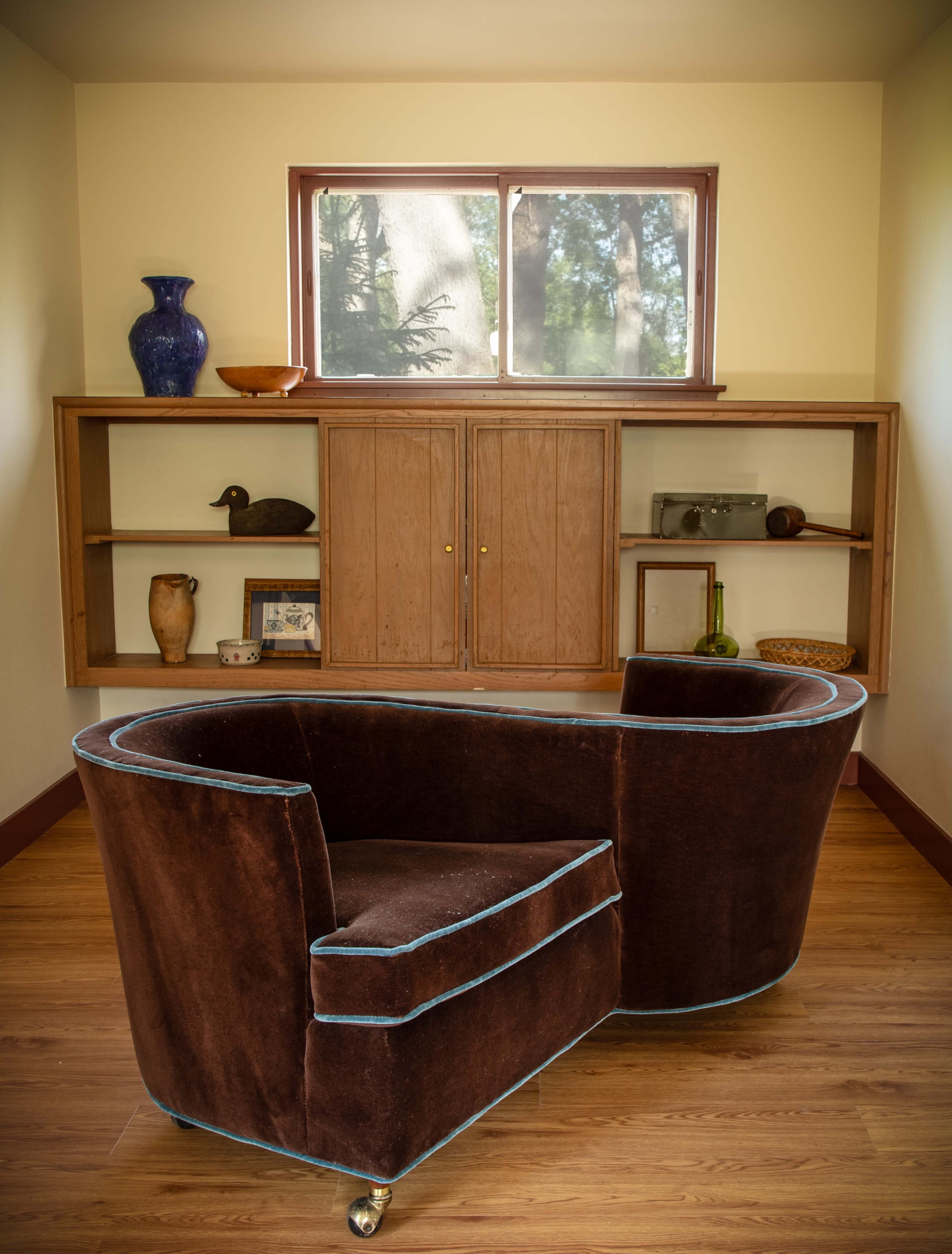 A plush, brown armchair with blue trim is positioned in front of a wooden cabinet and a window that lets in natural light.